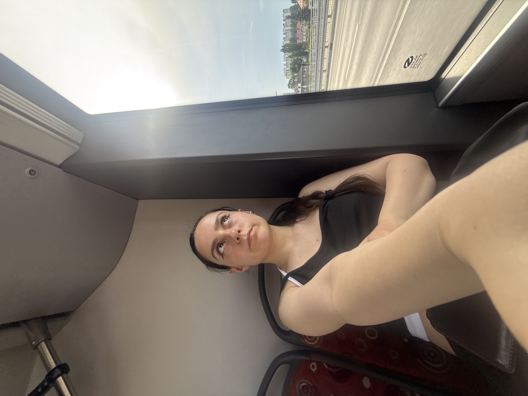 A young woman with dark hair and winged eyeliner is seated inside a bus, looking upwards and to her left. She is wearing a black tank top with white trim. Her left arm is bent across her chest, with her forearm extended forward. The bus interior is visible, including a red seat with a circular pattern behind her. To her right, a large window looks out onto a street with buildings and trees in the background. The sky is bright and hazy. The bus is likely on a route in Opole, Poland, possibly crossing a bridge as indicated by the road visible through the window.