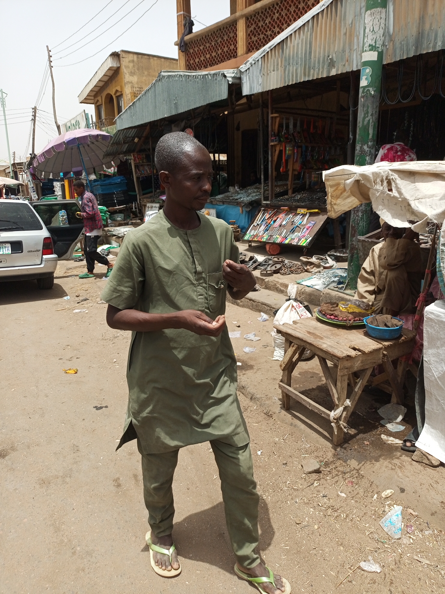 The scene is a street in Arawa, Nigeria. A man in an olive green tunic and pants stands on the dirt road. He wears sandals and holds something small in his hands. To his right, a wooden table displays food items, including fried snacks, a round container, and a plate of what appears to be grilled plantains. Behind the table, an individual sits under a partially draped covering. A market stall is visible in the background, displaying various tools and hardware. A person with a purple umbrella exits a silver car on the left. The area appears somewhat littered. Buildings with corrugated iron roofs and awnings line the street. Electrical wires stretch overhead.