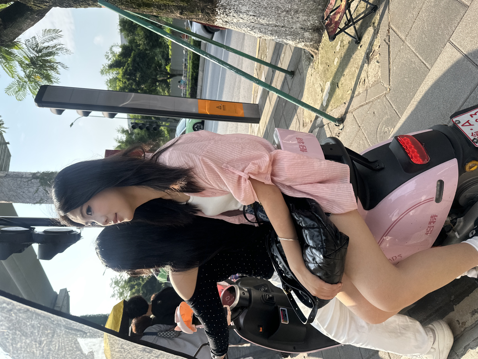 In Chongqing High-tech Zone, China, two young women are seated on a pink electric scooter. The woman in the foreground is wearing a pink, off-the-shoulder, striped shirt and white shorts. She has her left arm resting on a black quilted handbag. The second woman, seated behind her, wears a black off-the-shoulder top with rhinestones and white pants. They are on a sidewalk next to a street with traffic. A traffic light is visible in the background, along with trees and buildings. The lighting suggests it is daytime.