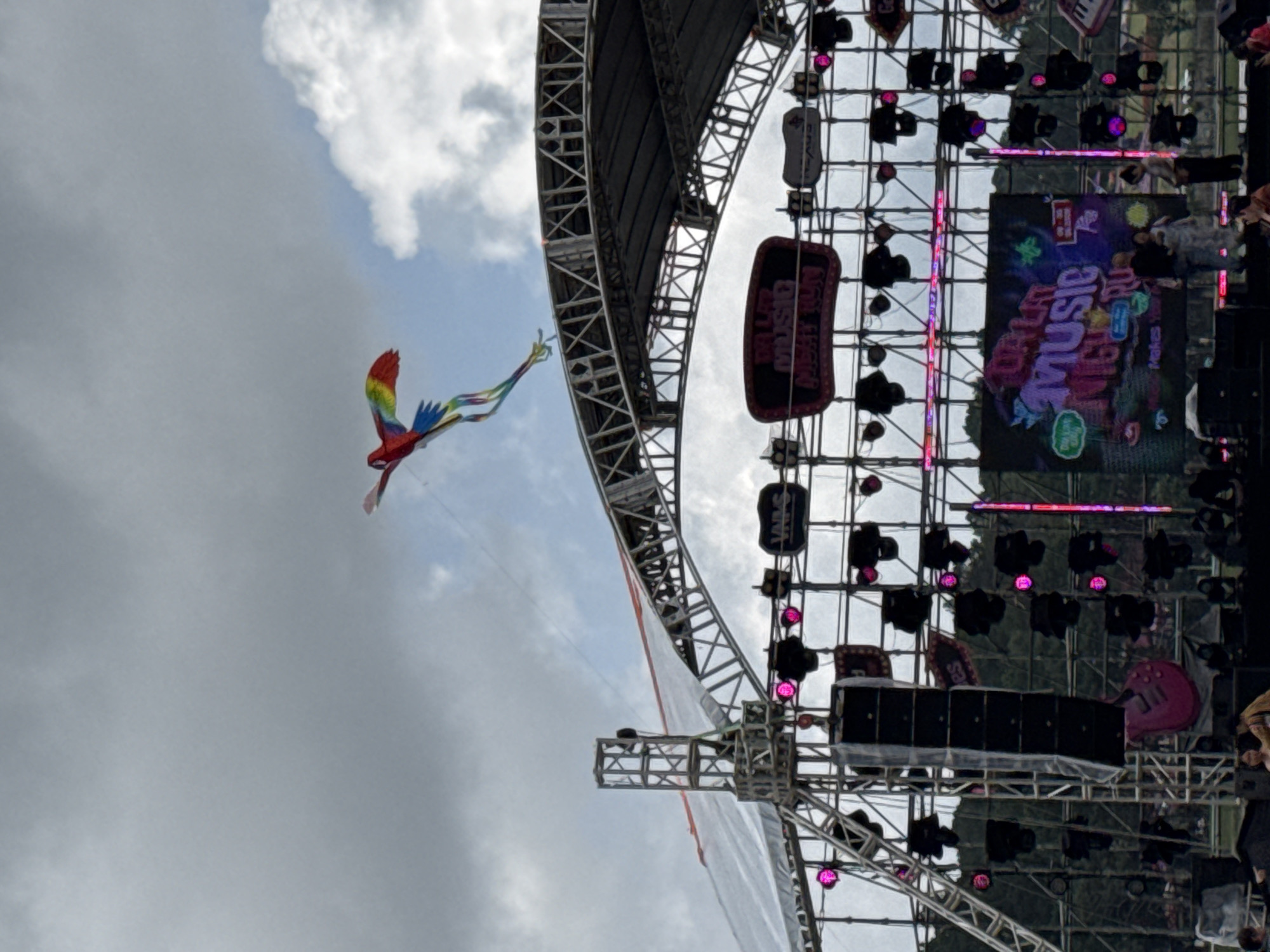 This image captures a vibrant outdoor scene, likely at a music festival or concert in Da Lat, Vietnam. The composition is dominated by a massive outdoor stage structure, constructed from intricate metal trusses, occupying the right side of the frame. This structure curves upwards, supporting numerous large speakers and lighting fixtures that emit a magenta or pink glow.
A large LED screen on the stage displays colorful graphics, prominently featuring the word "MUSIC" in a stylized font, along with other text that appears to say "DISCO" and possibly "FESTIVAL" or "DREAM," indicating the nature of the event. Several dark banners hanging from the stage structure also display text, including "DISCO MUSIC FESTIVAL" and "VANS." Visible behind the stage structure are hints of lush green foliage.
In striking contrast, against a partly cloudy sky that fills the left and upper portions of the image, a vivid, rainbow-colored kite soars gracefully. The kite is shaped like a large parrot or macaw, complete with a long, flowing multi-colored tail, adding a whimsical and festive element to the scene.
The weather appears to be overcast but bright, suggesting daytime. The overall impression is one of anticipation for a high-energy music event, balanced by the playful presence of the kite in the sky.