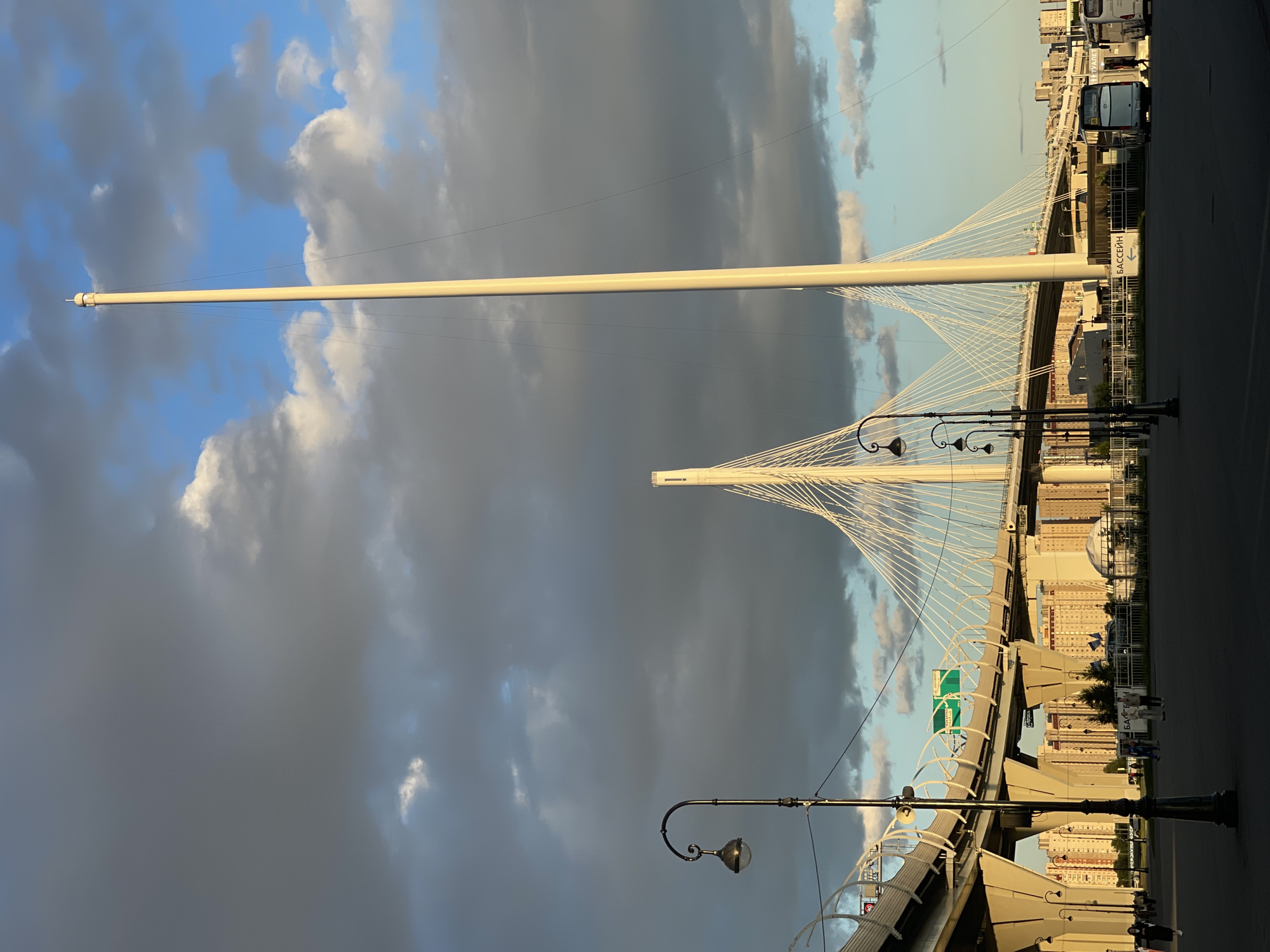 In an urban outdoor scene, a cable-stayed bridge dominates the background under a dramatic sky with a mix of dark clouds and patches of blue. The bridge features two tall, white pylons supporting a network of white cables. In the foreground, a dark lamppost with multiple arms and lights stands to the left. Beyond it, a portion of an elevated roadway with concrete supports is visible. A row of light-colored apartment buildings stretches across the mid-ground. On the paved ground in front of the buildings, a few people are walking. Two buses are parked to the right of the frame, one white and one light blue. Signage with Cyrillic text is visible on the buildings and near the buses. The lighting suggests it is either early morning or late afternoon.