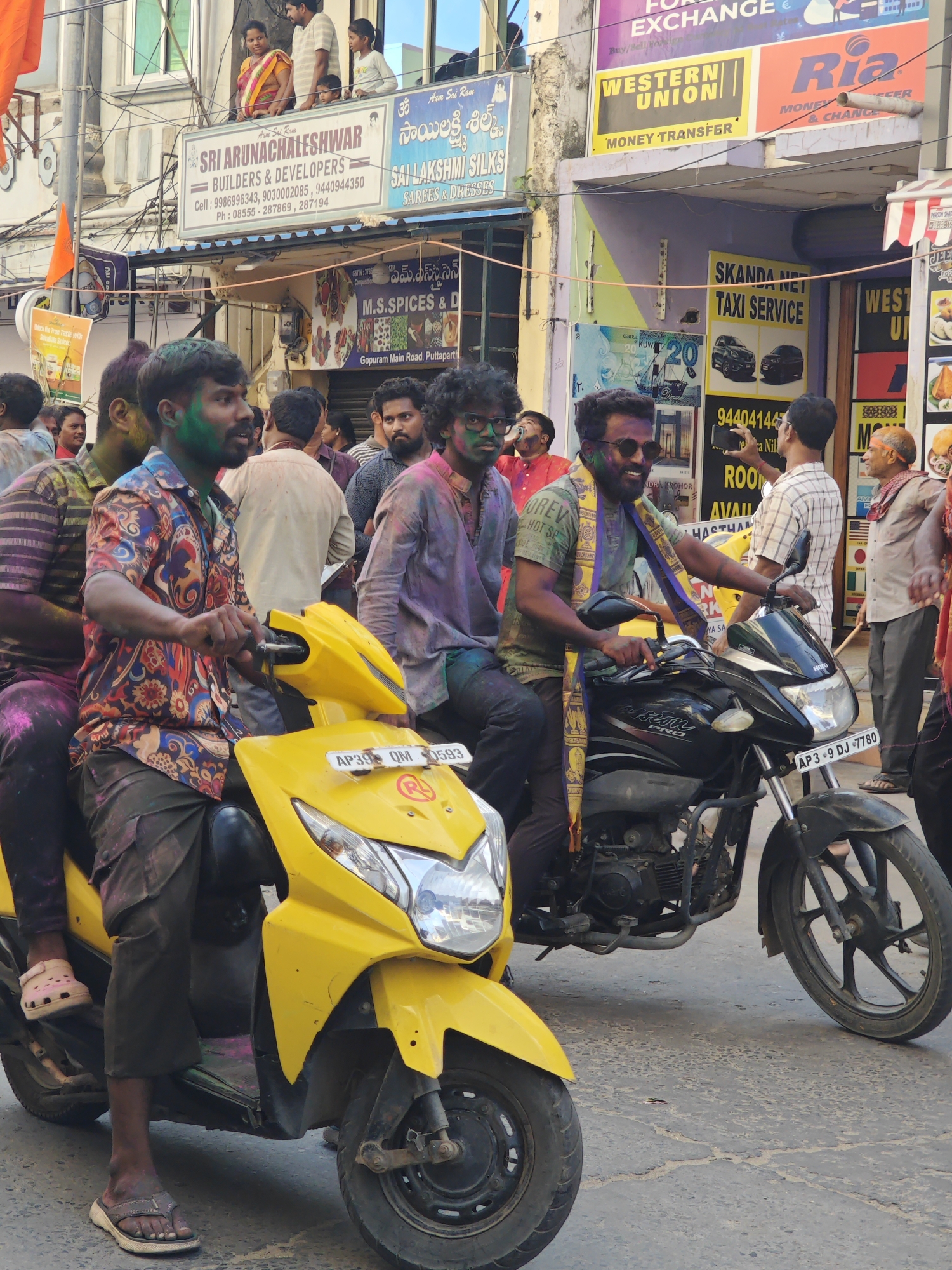 In Puttaparthi, India, on a street during the Holi festival, two men are riding on a yellow scooter and a black motorcycle. The man on the scooter, wearing a colorful shirt, has green powder on his face. The man on the motorcycle, wearing sunglasses and a yellow scarf, also has colored powder on his face. Both men are smiling. In the background, several other people are visible, some with colored powder on their faces and clothes. Buildings line the street, with signs indicating businesses such as "Sri Arunachaleshwar Builders & Developers" and "Sai Lakshmi Silks." The overall atmosphere suggests a festive celebration.