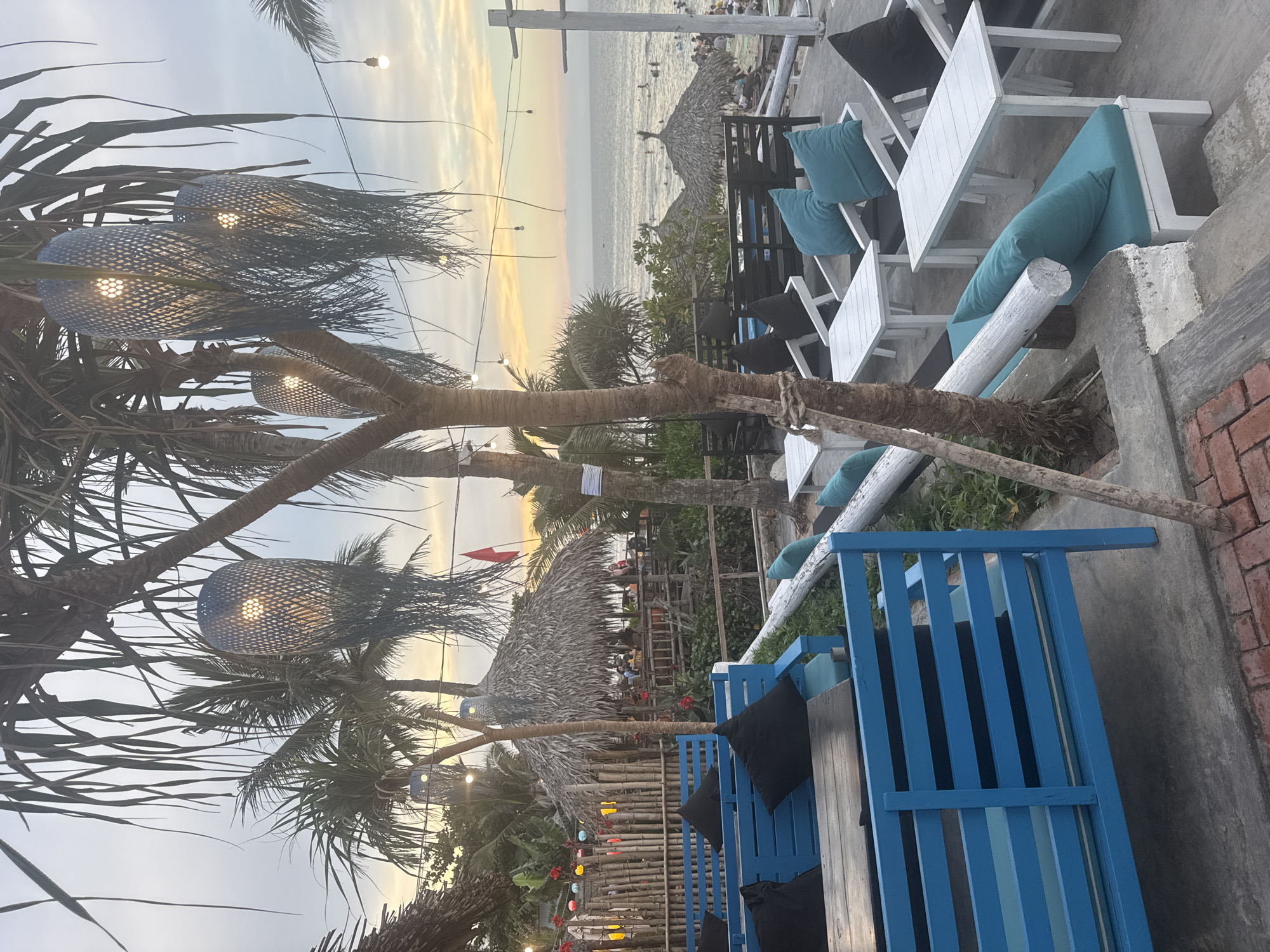 This exterior shot depicts an outdoor seating area in an unidentified location, possibly a city due to the presence of man-made structures. The sky is partly obscured by trees, and the time of day appears to be either early morning or late afternoon, based on the soft lighting. In the foreground, there is blue wooden seating with black cushions. Adjacent to this are white tables and chairs with blue cushions. Beyond the seating area, there is a partial view of what appears to be a beach or coastline. There are people visible in the distance on the beach, suggesting an active environment. The trees in the scene have string lights hanging from them, adding to the ambiance. The overall scene suggests a relaxed, beachside setting.