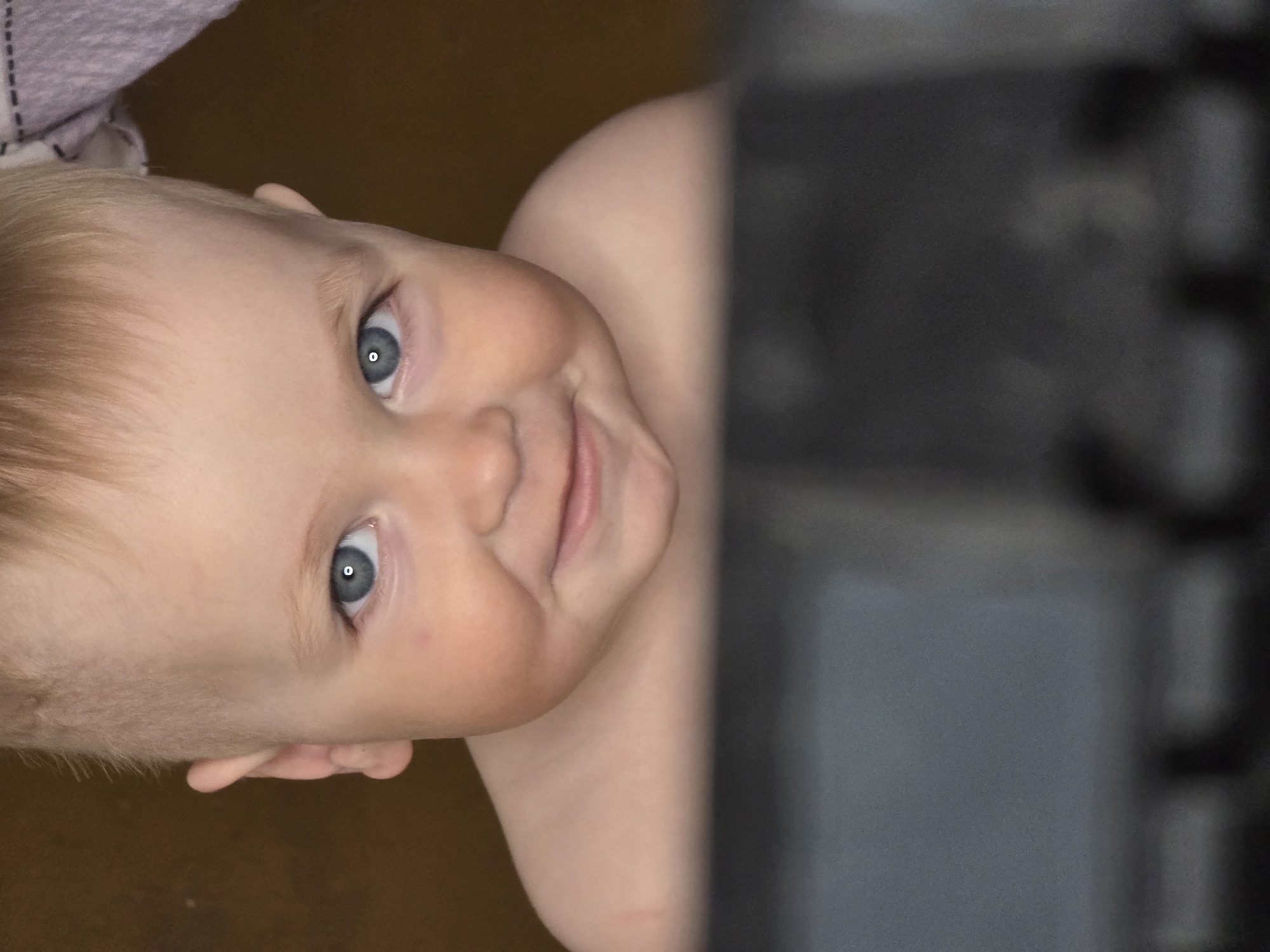A portrait-style photograph captures the engaging face of a baby or young toddler. The child, with light blonde hair and bright blue-grey eyes, is seen from a tilted perspective, appearing to look upwards or sideways. The baby has fair skin and plump, rosy cheeks, offering a gentle, endearing smile directly towards the viewer. The gaze is direct and curious, with a visible reflection of a light source in the pupils.
Part of the baby's bare shoulder and arm are visible, suggesting they might be dressed in a sleeveless garment or partially uncovered. In the top left corner, a piece of light-colored, possibly knitted, fabric is visible. The bottom left shows a dark, warm-toned surface, likely a wooden floor. The right side of the image is dominated by a dark, heavily blurred vertical object, possibly furniture or a doorway, which creates a strong contrast with the baby's bright face.
The overall impression is one of warmth and innocence, with the baby's gentle and happy expression being the central focus. The image appears to be taken indoors, likely in a home setting. This scene is contextualized as being in Chaikovsky, Russia. There is no visible text in the image.