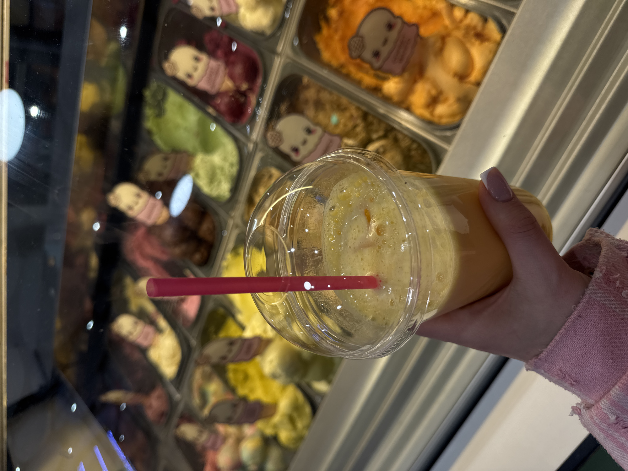 An individual is holding a plastic cup with a red straw in front of a glass display case filled with various flavors of gelato. The cup contains a yellow liquid, possibly a smoothie. The gelato flavors in the display case include pink, yellow, green, brown, and orange. Some gelato containers have cartoon character stickers. The location appears to be a gelato shop. The specific location in Moscow, Russia, cannot be determined from the image.