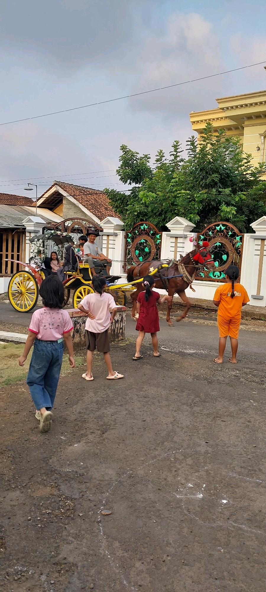 In Keling, Indonesia, a horse-drawn carriage moves down a paved street. The carriage, yellow with decorative embellishments, carries at least three passengers and a driver. A brown horse pulls the carriage. Several children are visible on the street, some walking and others standing. One child in a pink shirt and blue jeans walks away from the camera. Another child in a pink shirt and brown shorts stands with hands on hips. A child in a red dress stands facing the carriage. A child in an orange shirt and shorts stands facing away from the carriage. Behind the carriage, a decorative fence and trees are visible, with a building in the background. The sky is overcast.