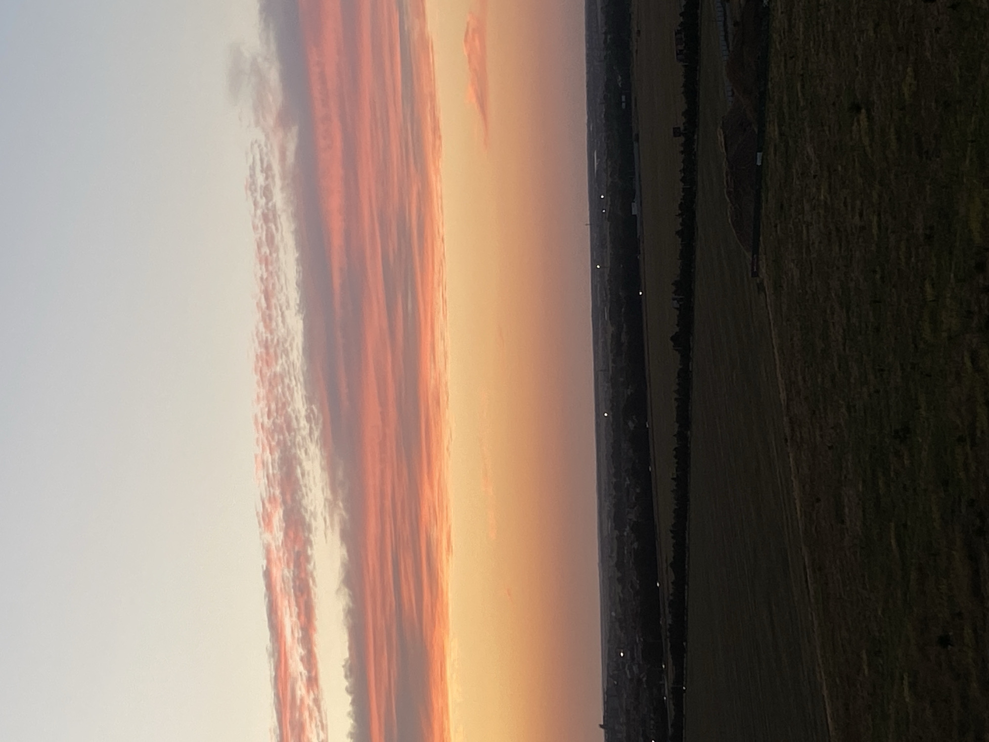 This is a wide-angle, eye-level outdoor photograph taken at dusk in the city of Chornomorsk, Ukraine. The sky is filled with streaks of pink and orange clouds, transitioning to a pale blue at the top of the frame. Below the sky, a dark, flat landscape is visible, featuring agricultural fields and a distant treeline. The fields appear to be cultivated, with some areas showing signs of recent harvest or storage. Several lights are visible in the distance, suggesting the presence of a settlement or infrastructure. The overall impression is one of a serene, rural sunset scene on the outskirts of a city.