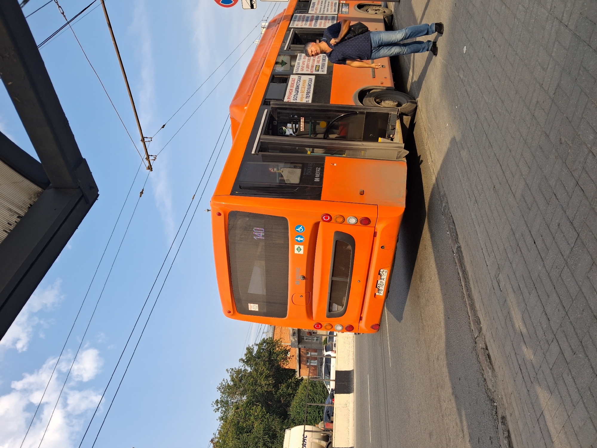 An orange NEFAZ bus with the route number 140 displayed on its rear is parked at a bus stop on a sunny day in Novosibirsk, Russia. A man wearing a dark blue polo shirt and jeans stands near the open doors of the bus, carrying a dark backpack over his right shoulder. The bus stop has a paved waiting area and is located next to a road with trolleybus wires overhead. In the background, a brick building with the word "ТЕАТР" (Theater) visible on its facade is partially obscured by trees. The license plate of the bus is PB18PA 154.