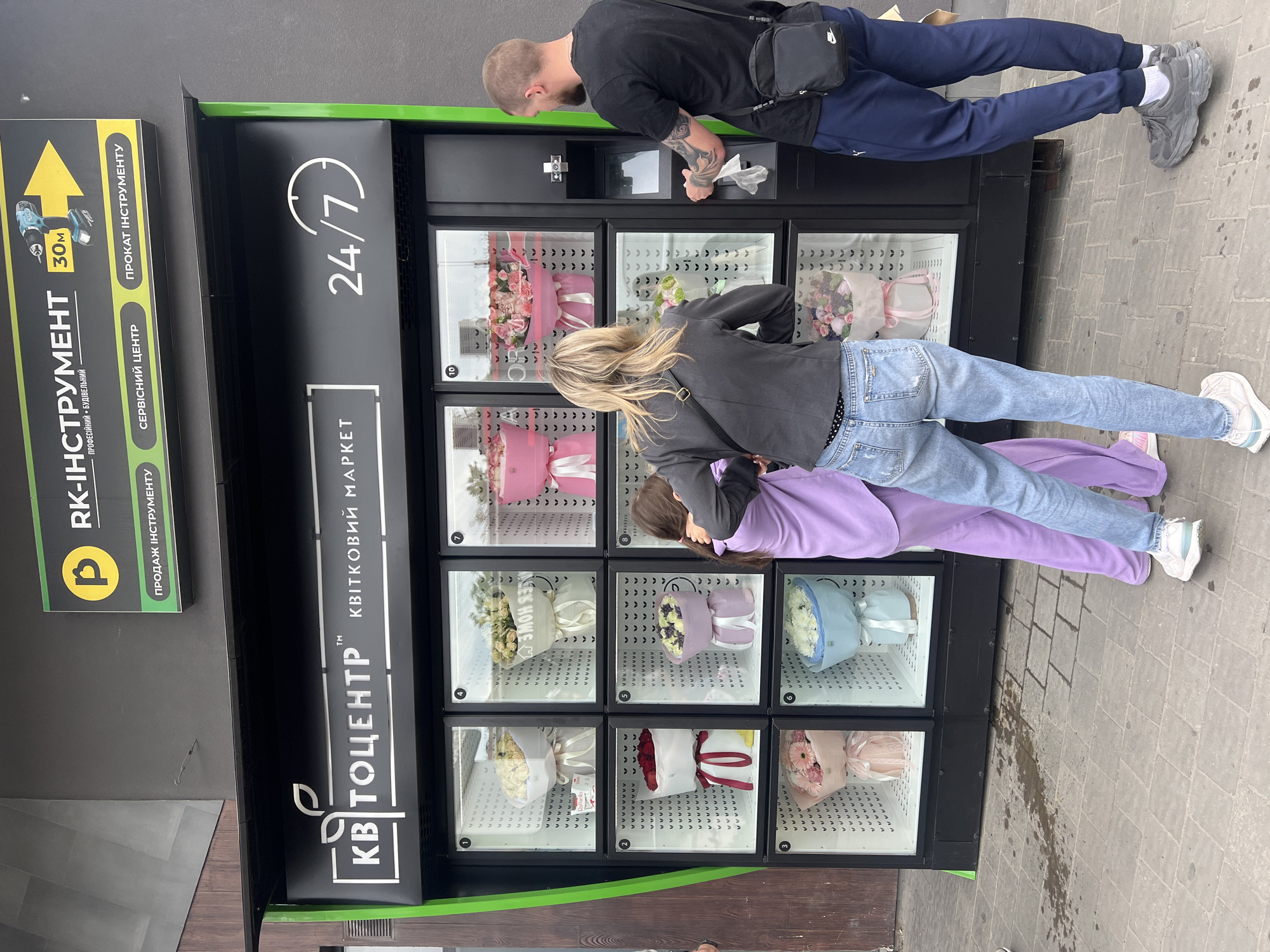 The image depicts a flower vending machine in what is identified from other signage as the city of Ternopil, Ukraine. Three individuals are interacting with the machine: a man is at the top dispensing area, a woman is in the middle, and a child is positioned below her, looking at the flowers on display. The vending machine, labeled "KVITO CENTER," operates "24/7." It displays a variety of bouquets in individual compartments. A sign next to the flower vending machine advertises "RK-INSTRUMENT," offering tool sales, service, and rentals, indicated by a yellow arrow and a graphic of a power drill. The environment consists of a sidewalk with patterned paving stones and a dark-colored exterior wall.
