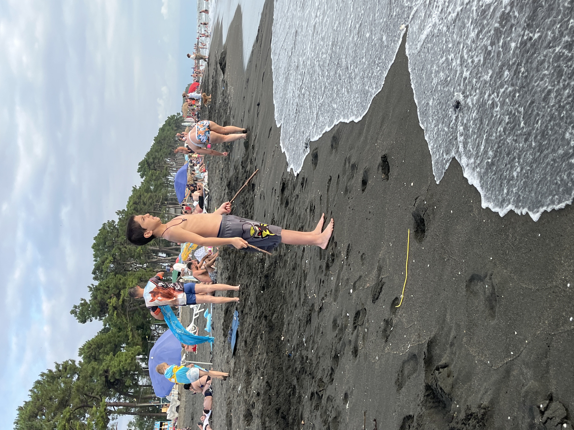 This is a medium shot, eye-level, outdoor photograph taken on a black sand beach in Ureki, Georgia. The sky is overcast with scattered clouds. In the foreground, a young boy, appearing to be around 8-10 years old, stands facing right, holding a thin stick. He is wearing grey patterned shorts. His feet are bare on the dark sand. The sand is wet near the water's edge, with small white shells scattered across it. Footprints are visible in the sand. The ocean is on the right side of the frame, with waves gently lapping the shore. In the mid-ground, to the left of the boy, another young person in a blue towel is visible. Further back, a crowd of people are scattered on the beach, some sitting under blue umbrellas, and others walking or swimming in the ocean. Pine trees line the background of the beach, with a hazy shoreline beyond.