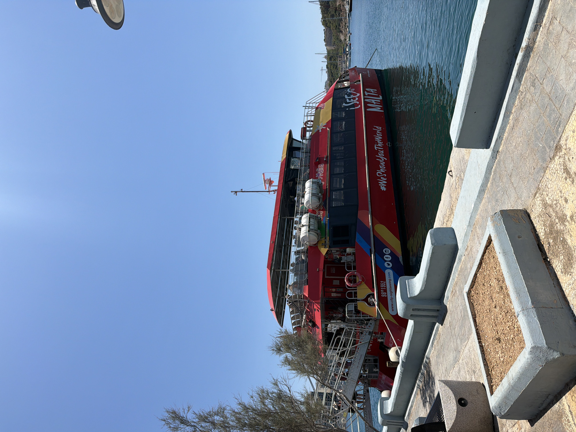 A vibrant red and yellow passenger ferry, identified as "SEE MALTA" with the hashtag "#WeShowYouTheWorld", is docked at a waterfront promenade in Sliema, Malta. The ferry, adorned with rainbow-colored stripes along its hull, is primarily red with a yellow upper deck and canopy. Several passengers are visible on the upper deck, seated and enjoying the sunny weather. The ferry's name is prominently displayed in large white lettering on its side, with smaller text indicating "CITY SIGHTSEEING.COM" and social media icons.
The promenade in the foreground is paved with interlocking bricks and features several concrete planters, one of which contains dry soil and small stones. A grey concrete barrier runs along the edge of the promenade, separating it from the calm, blue water. In the background, other boats and masts of sailboats are visible in the harbor, and a hilly coastline with buildings can be seen under a clear blue sky. The overall scene depicts a typical day at a popular tourist destination in Sliema.