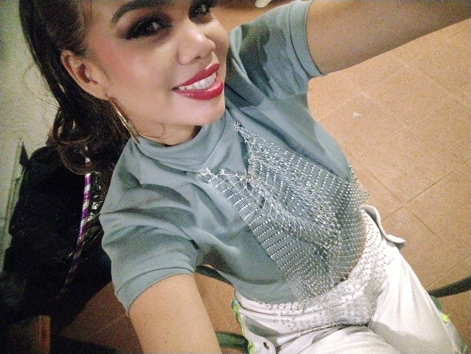 A woman poses for a selfie indoors. She has dark hair pulled back, dark eye makeup, and bright red lipstick. She is wearing a light grey, short-sleeved turtleneck shirt adorned with a decorative silver mesh design on the front. She is also wearing white pants with a decorative belt. A black object is partially visible in the lower left corner. The background is out of focus, showing parts of a wall and floor. It is not possible to determine the location in the city of Porlamar, Venezuela, from this image.