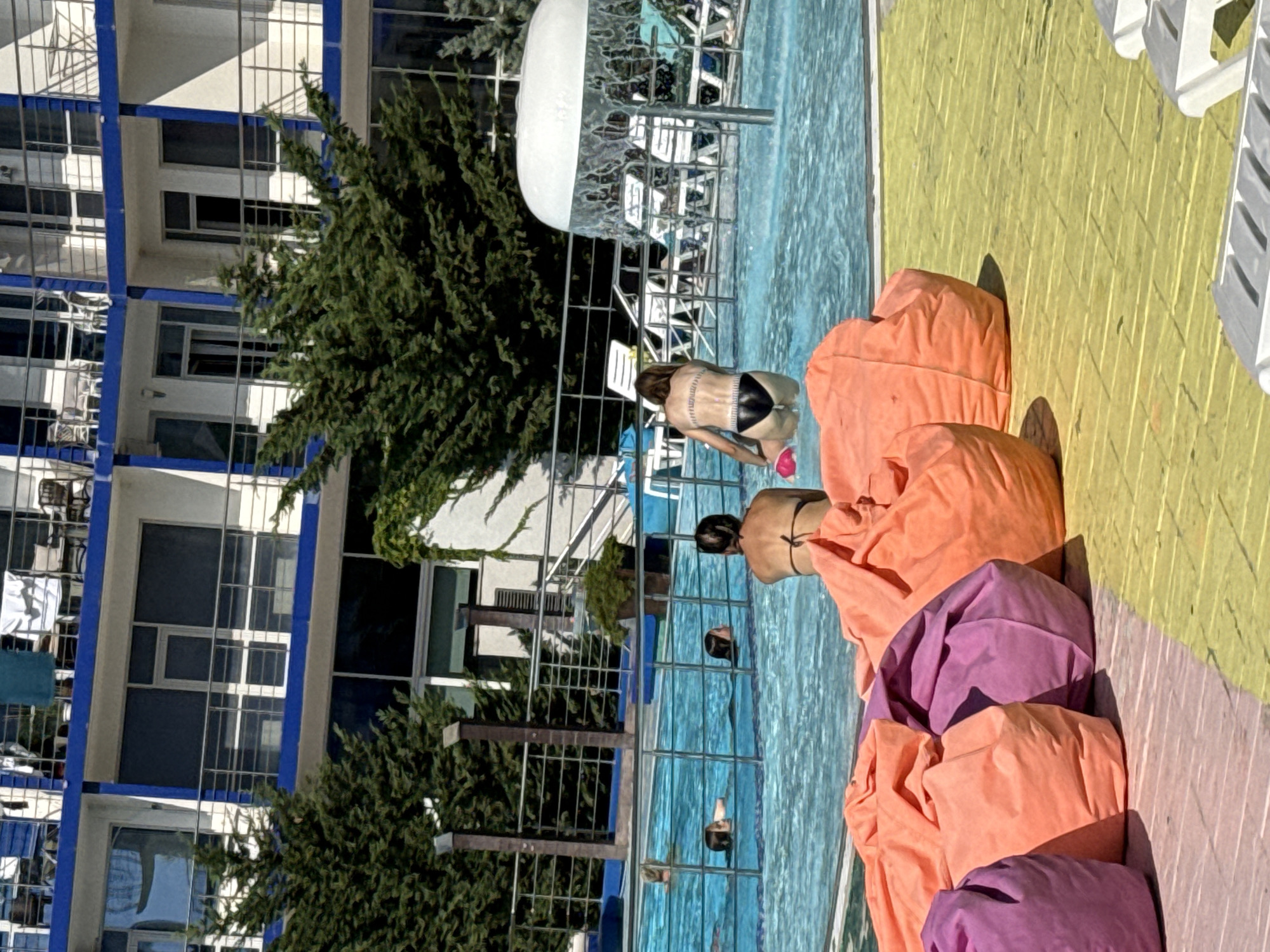 The image depicts an outdoor swimming pool area at a resort located in Sevastopol, Ukraine. In the foreground, several large, colorful beanbag chairs are arranged on a yellow-tiled deck. The swimming pool itself is visible in the middle ground, filled with clear blue water. Several people are in the pool, including one individual partially submerged and another sitting on the edge. A woman in a black bikini is seen standing in the pool, facing away from the camera, near a mushroom-shaped water feature. In the background, a multi-story building with blue trim and balconies, indicative of hotel or resort accommodations, is visible, surrounded by lush green trees. The overall scene suggests a warm, sunny day at a recreational facility.