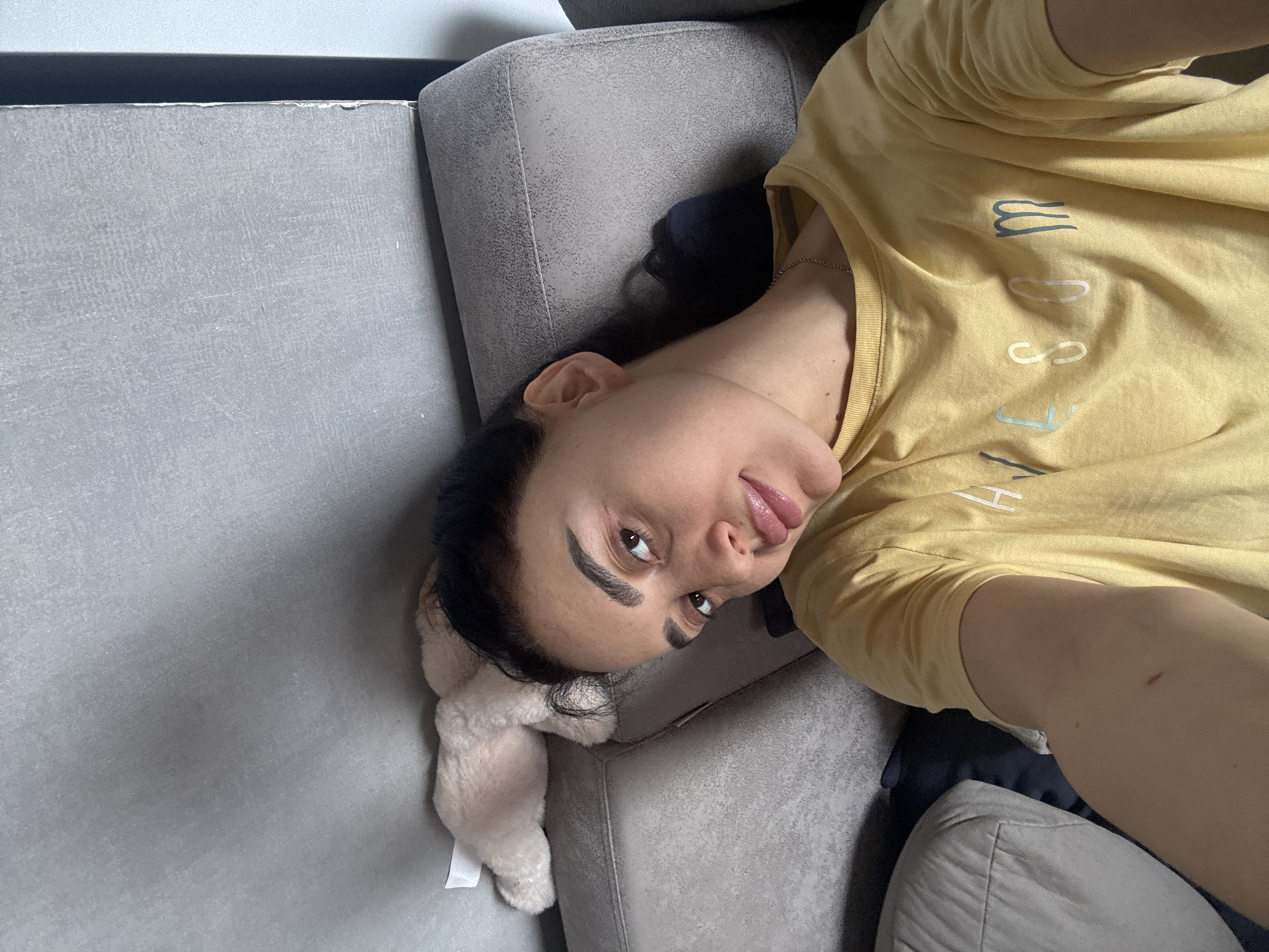 A woman is lying on a gray couch. She is wearing a yellow t-shirt. A stuffed animal is near her head. The location of the photo is unknown, and there is no evidence to suggest that it was taken in Tbilisi, Georgia.