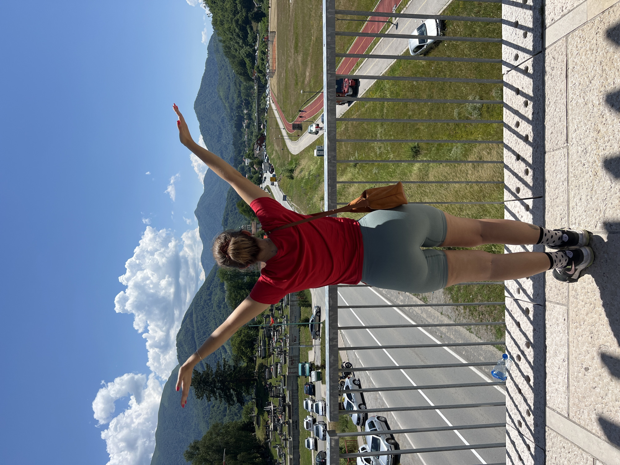 In Mojkovac, Montenegro, a young woman stands on a bridge overlooking a scenic landscape. She is viewed from behind, with her arms outstretched towards the sky, embracing the sunny day. She wears a vibrant red t-shirt and light green shorts, complemented by sneakers and black socks with white polka dots. A brown crossbody bag is slung over her right shoulder.
The view from the bridge reveals a road winding through the town, with cars and a truck driving along it. Beyond the road, a sports field with a red track is visible, and further in the distance, verdant mountains rise against a clear blue sky dotted with fluffy white clouds. A cemetery with numerous gravestones can be seen on a hill to the left. The bridge railing in the foreground casts a shadow on the stone walkway.