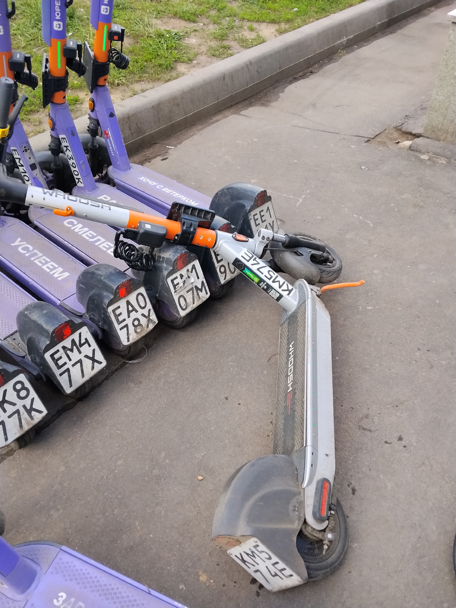 The image shows a row of parked electric scooters in Kotelniki, Russia. The scooters are primarily purple and grey, with branding from companies like "Whoosh" and "ЮРЕНТ". Each scooter has a license plate, with visible plates including "EM4 77X", "EAO 78X", "EM1 07M", "EE1 AY", and "KM5 74E". Some scooters are closely packed together while one grey scooter is lying on its side. The scooters are parked on a paved surface, likely a sidewalk or parking area, next to a curb. The surrounding environment appears to be an urban area with grass visible in the background.