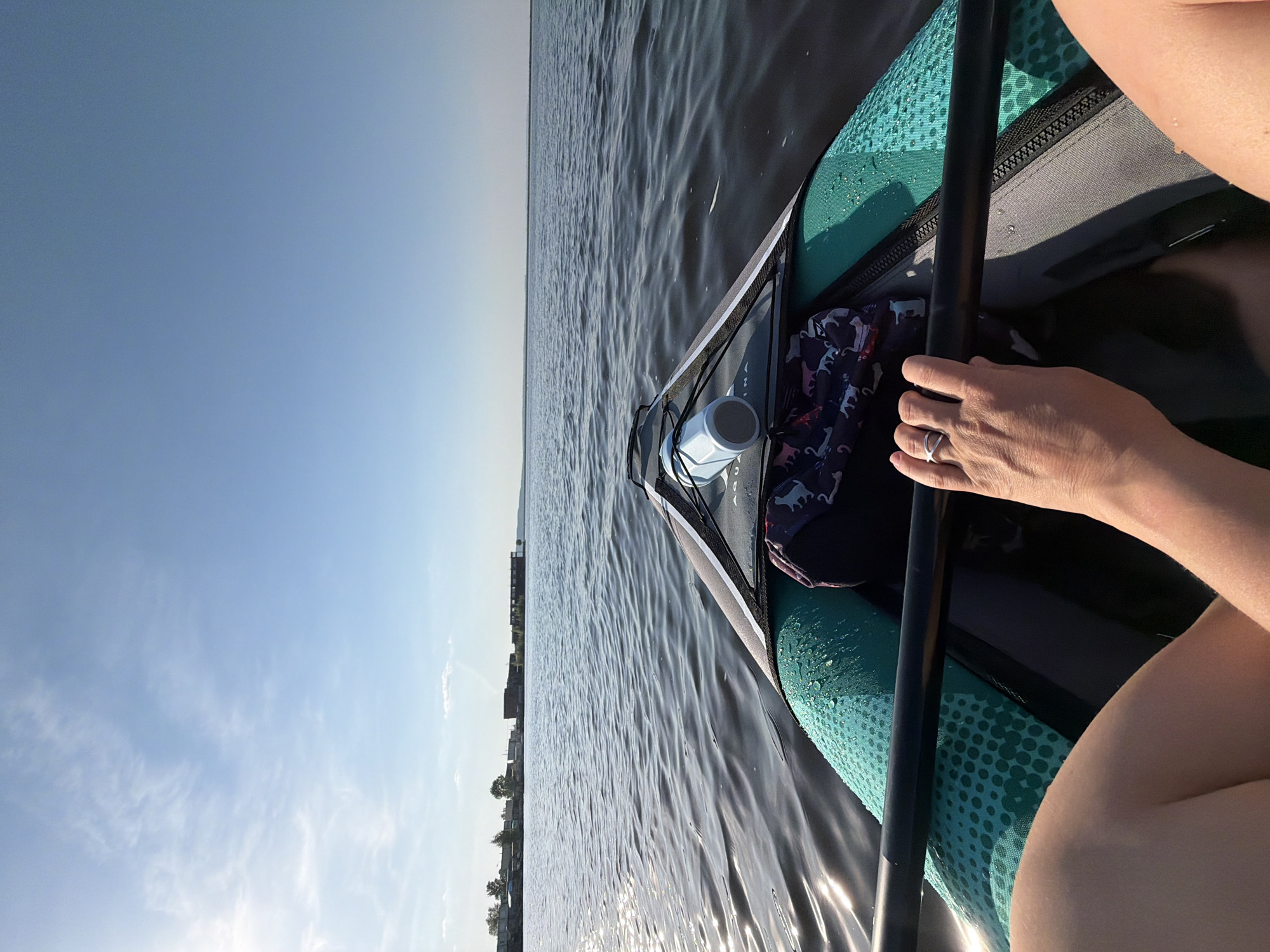 A first-person perspective photograph from a water vessel, likely a kayak or inflatable boat, shows a partially visible person holding a paddle with their left hand. The water stretches towards the horizon under a clear, light blue sky, with a hint of land visible in the distance to the left. The vessel's bow has a storage area with a water bottle and a patterned bag inside. The location is likely in or near Chaikovsky, Russia, given the query context.