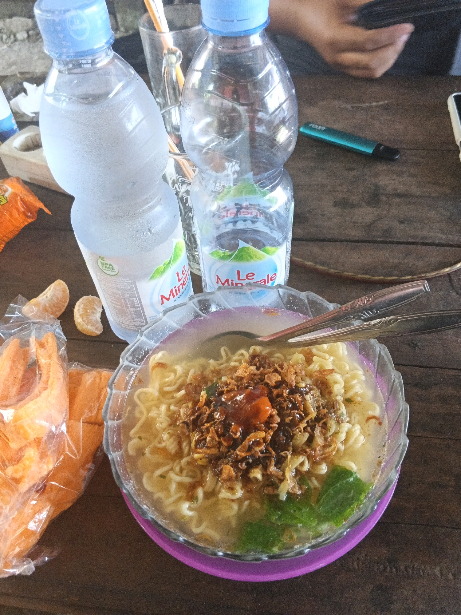On a weathered, wooden table, a bowl of instant noodles, topped with fried shallots and a dollop of red sauce, sits on a purple plate. Beside it, two bottles of "Le Minerale" bottled water, one condensation-covered and the other clear, are present. A bag of orange crackers, along with two slices of tangerine, are also visible. In the background, a hand is visible holding a black phone, and a teal colored vape pen lies on the table. The scene appears to be set outdoors, possibly at a roadside eatery in Kecamatan Pati, Indonesia, given the rustic table and casual arrangement of food and drinks. The lighting suggests it is daytime, and the overall atmosphere is informal and relaxed.