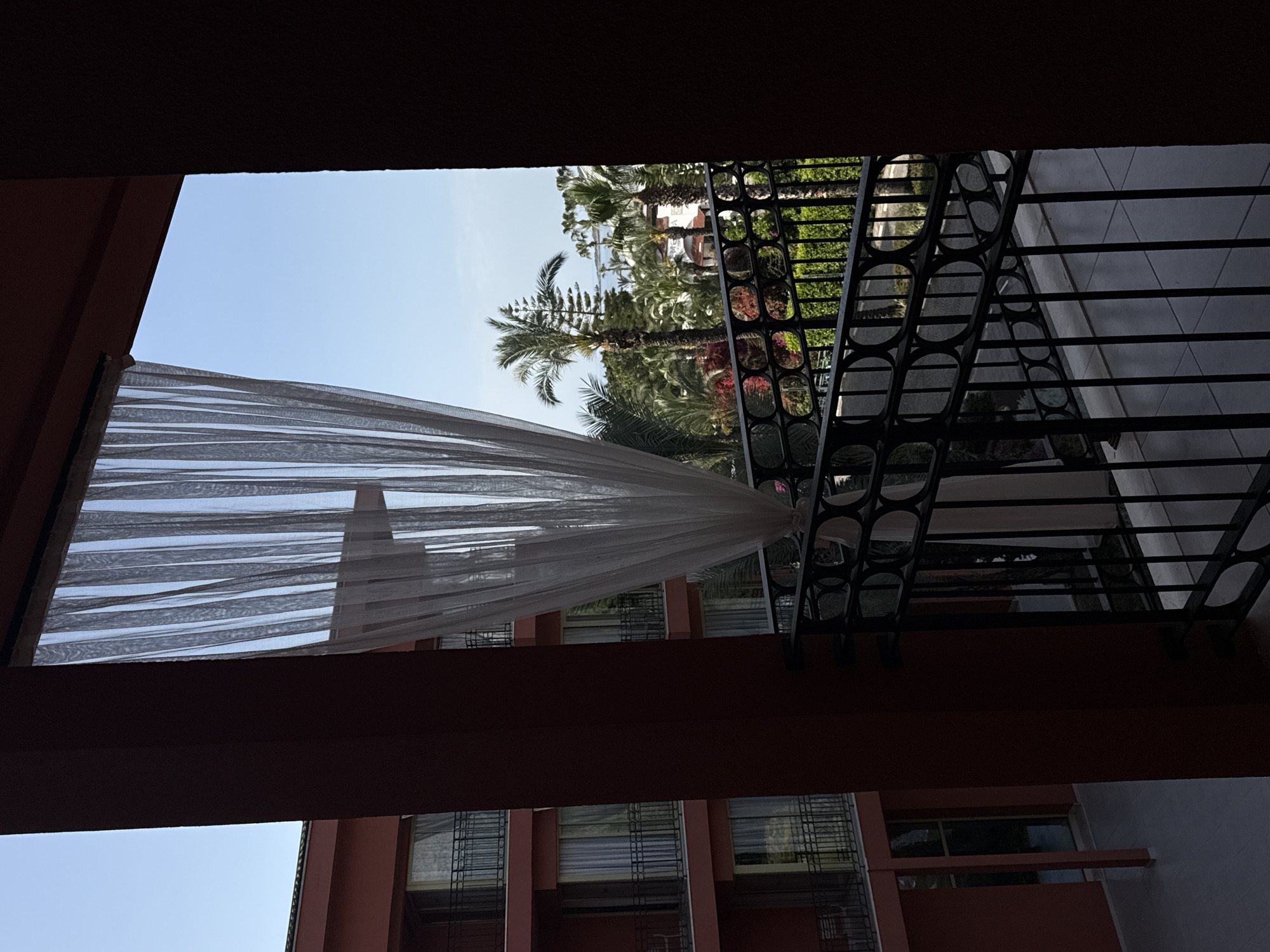 The image shows a view from a balcony, likely in Kemer, Turkey, based on the presence of palm trees and the architecture. The balcony has a black metal railing with a decorative circular pattern. A sheer white curtain is draped from the balcony above, obscuring the view slightly. The view beyond the balcony includes palm trees, greenery, and parts of a building. The building appears to be terracotta-colored with multiple windows, suggesting it could be a hotel or apartment complex. The sky is visible in the upper portion of the frame. The overall scene has a relaxed, vacation-like atmosphere.