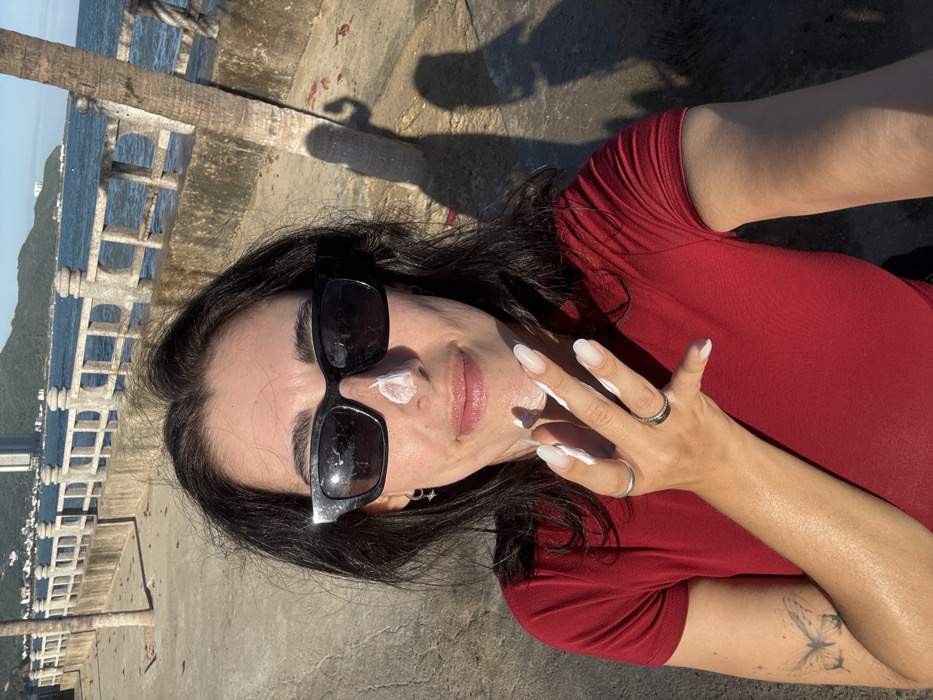 An image captures a female individual at a coastal location identified as Nha Trang, Vietnam. The subject, positioned centrally, has dark hair, wears black rectangular sunglasses, and a red short-sleeved shirt. A dollop of white sunscreen is visible on the bridge of her nose, and her left hand, adorned with white-tipped manicured fingernails, holds additional sunscreen on her fingertips, positioned near her face. Rings are present on two fingers of her left hand, and a butterfly tattoo is visible on her left forearm.
The background reveals a concrete promenade or seawall with decorative balusters separating a paved walkway from a body of water. A segment of a palm tree trunk is visible on the right, casting a distinct shadow on the sun-drenched concrete ground. Beyond the seawall, calm blue water extends to a distant coastline featuring green hills and several tall, multi-story buildings. The sky above is clear. Strong sunlight is evident, creating well-defined shadows on the ground.
