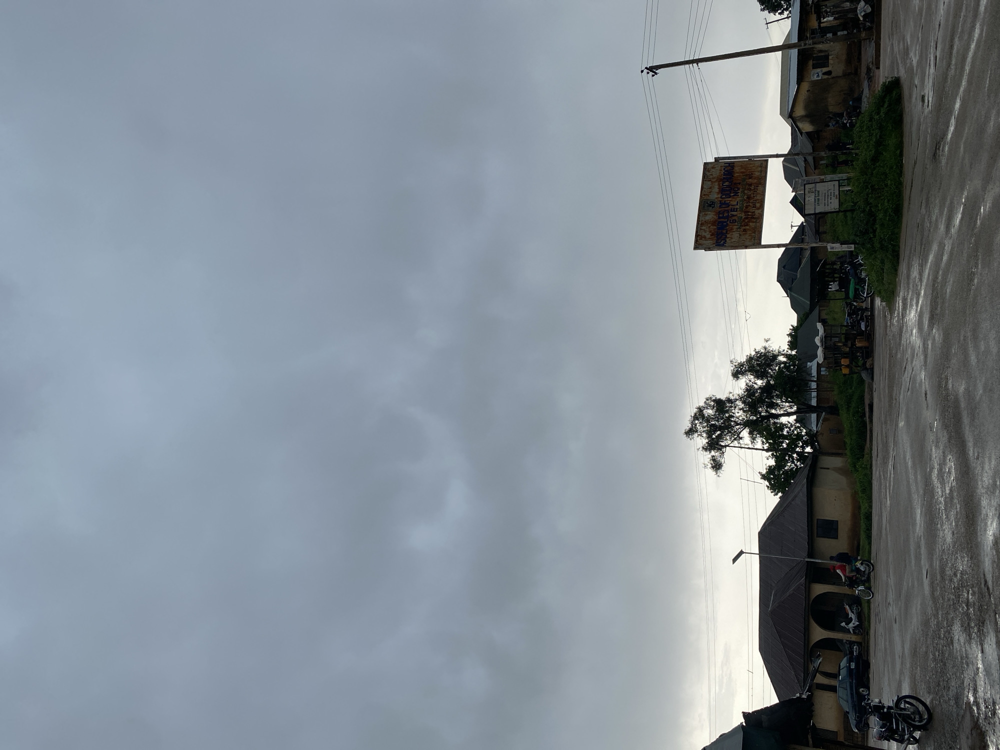 This media file captures a scene in Barikin Gidar Kanat, Nigeria. The image is oriented vertically, with the sky dominating the left side and a street scene on the right. The sky is overcast with gray clouds, suggesting a gloomy or rainy day. The ground appears wet, with reflections indicating recent rainfall.
On the right side of the image, there is a street lined with buildings and vegetation. Several motorcycles are parked along the street and in front of the buildings, suggesting local transportation. A few people are visible, some on or near the motorcycles. One person is wearing a red shirt. A car is also parked on the street.
A prominent feature is a large, weathered sign with visible text. The text, though partially obscured and faded, appears to read "ASSEMBLIES OF GOD CHURCH" and "GIKEL KGUN-CHURCH". Below this, there is more text, including "BUHL STATE". Another smaller sign is also visible near the main sign.
The architecture of the buildings suggests a local style. A large tree with dense foliage is present, with its branches reaching towards the sky. Electrical poles and wires are also visible, indicating infrastructure in the area. The overall mood is subdued, likely due to the overcast weather. The scene appears to be a typical street view in a populated area, possibly a town or village, with religious institutions and local businesses present. The time of day is unclear due to the cloud cover, but the light suggests it is daytime.