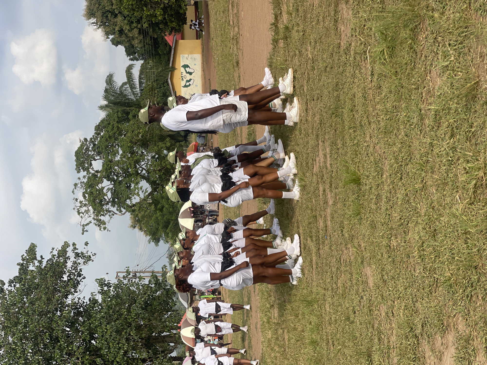 In Emure-Ekiti, Nigeria, a group of individuals, predominantly young women wearing white t-shirts, white shorts, green caps, and black fanny packs, are marching in formation on a grassy field under a cloudy sky. Some individuals are walking individually in the background, also wearing similar attire. The scene appears to be an outdoor event or training exercise, possibly related to a national youth service program given the uniforms. In the background, there are trees, tents, and a yellow building with green murals depicting a handshake and wings, and the word "UNITY." The ground is covered with dry grass and dirt.