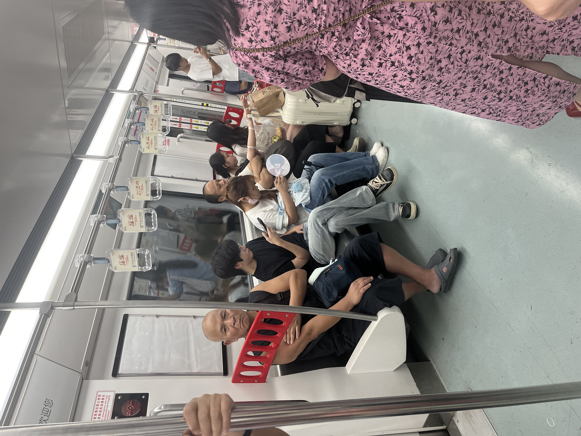 A group of people are traveling on a subway train in Jiulongpo District, China. The train interior is brightly lit with fluorescent lights. On the left, a bald man in a black sleeveless shirt and shorts sits with a red object resembling a handle or support propped on his arm. He is wearing grey slip-on shoes. In front of him, part of another passenger's arm is visible, holding onto a silver pole. Further down the carriage, several other passengers are seated and standing. One man in a white t-shirt is looking at his phone. Another passenger, a woman, is wearing a pink floral dress and is standing near the foreground, facing away from the camera. Luggage, including a white suitcase, is visible on the floor. Signs are posted on the train walls, some with Chinese characters. The floor is a speckled light blue.