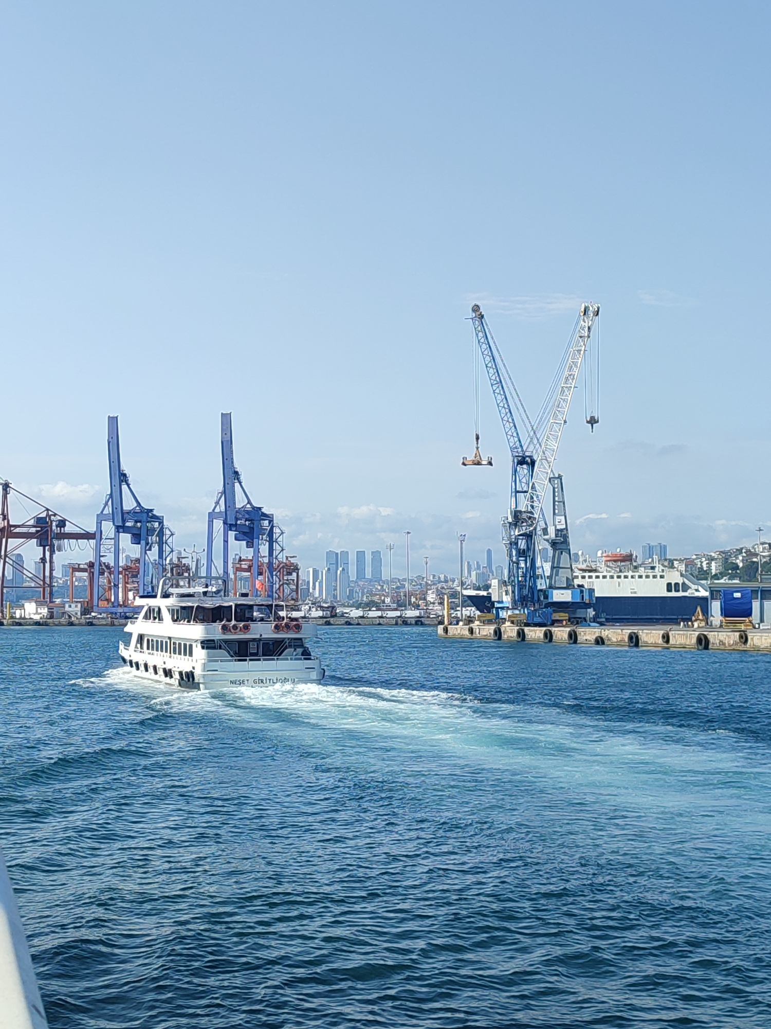 A ferry named "NESET GIRTUCIGLU" sails through the blue waters of the Bosphorus Strait in Istanbul, Turkey. The ferry, a white vessel with multiple windows and decks, is captured from behind, leaving a foamy wake. In the background, a bustling port area is visible, dominated by towering blue and red cranes, indicative of container handling operations. A large blue and white ship is docked at the pier. The cityscape of Istanbul forms a backdrop, with numerous buildings of varying heights clustered on a hillside. The sky is clear and blue with a few scattered clouds, suggesting a bright, sunny day. The overall scene depicts a vibrant maritime and urban environment.