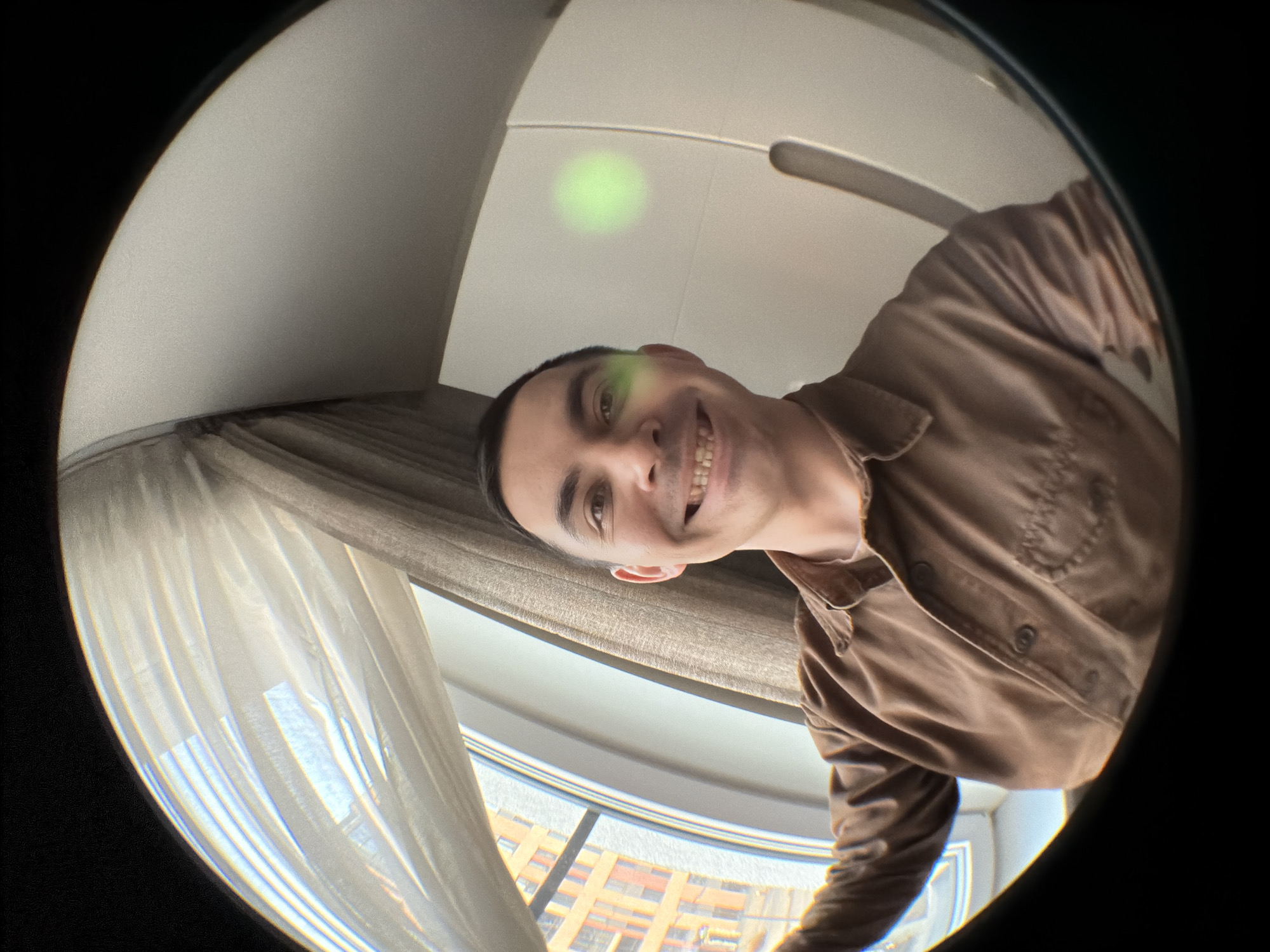 A man is holding the camera in a fisheye, close-up selfie, wearing a brown collared shirt. He is smiling. The background reveals a room with a window, curtains, and a partial view of buildings. The location in the city of Izhevsk, Russia is not clear from the image.