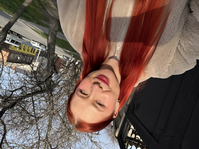A young adult female is depicted in a selfie, positioned outdoors on an urban street in Rostov-on-Don, Russia. The subject has long, straight red hair, a fair complexion with visible pink blush and lipstick, and wears a light beige zippered fleece jacket. Visible accessories include a septum nose ring and multiple ear piercings (two lobe, one helix) on the left ear. She is smiling slightly and looking towards the camera.
The background to the subject's immediate left features a dark metal gate with decorative elements, illuminated by strong sunlight. To her right, a street scene unfolds under a clear blue sky. Deciduous trees with bare branches line the street, indicating a season outside of summer. A paved road is visible with several vehicles, including a white sedan and a white van. Buildings line the street, notably one with a yellow lower level facade and a darker upper story. The overall lighting suggests a sunny day.