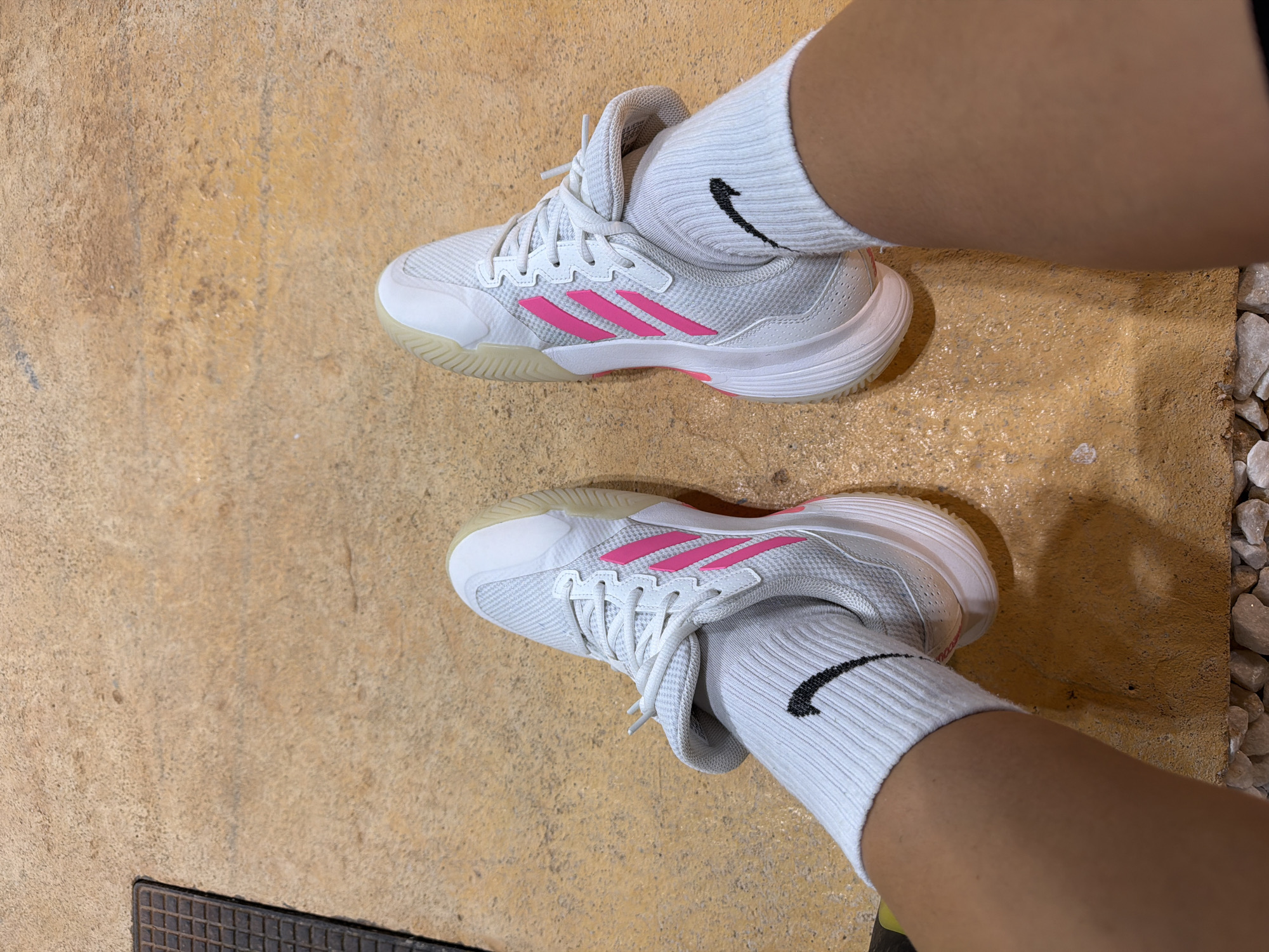 This is a top-down, medium shot photograph of a person's feet wearing white Nike socks and white Adidas athletic shoes with pink accents. The person is standing on a textured, yellowish-tan surface that appears to be concrete or asphalt. A dark, grated metal plate is visible in the upper left corner of the frame. The scene suggests an outdoor urban environment, potentially a walkway or court. The lighting is bright, casting shadows on the ground. This image was taken in Finestrat, Spain.