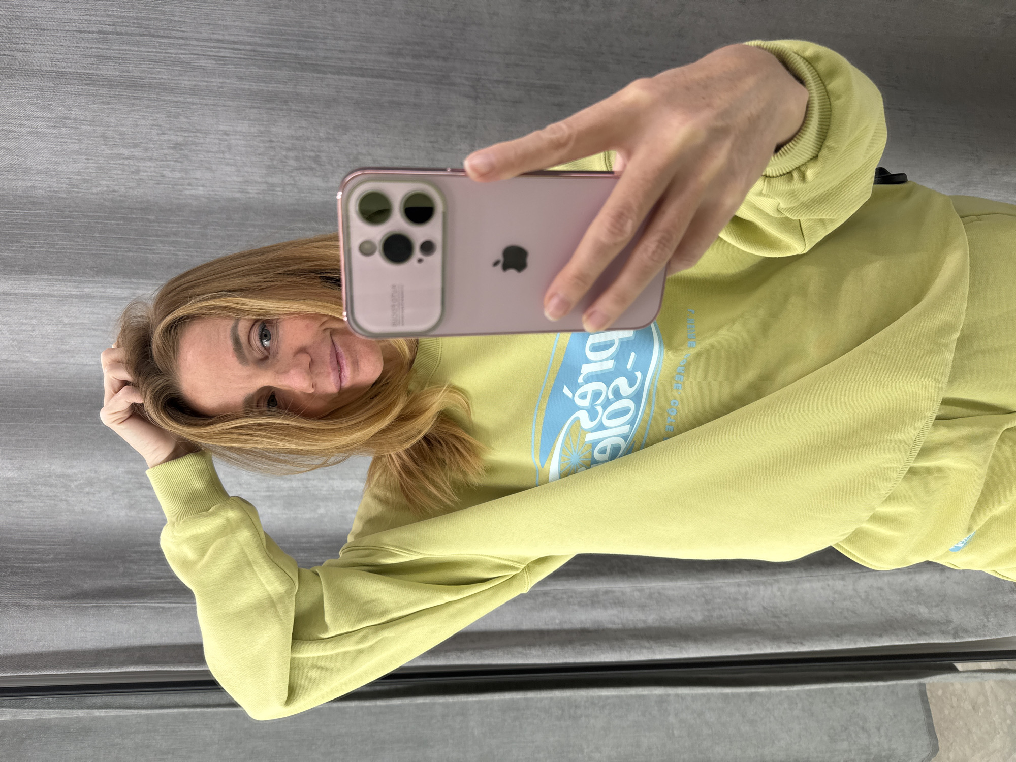 A woman is captured in a selfie, holding a pink iPhone with her right hand and looking directly into the camera with a pleasant, slight smile. Her long, light brown hair frames her face, with strands falling over her shoulders. Her left hand is raised, gently touching her hair near her head.
She is wearing a long-sleeved sweatshirt of a distinct lime green or chartreuse color. The front of the sweatshirt features a mirrored graphic design in light blue. Prominently visible within this design is the mirrored number "2016". Below this, mirrored text that appears to be "FREE COFFEE" is partially discernible, along with what could be "BIG" above the number, and a stylized wheel or sun motif. The sweatshirt has ribbed cuffs.
The iPhone, likely a Pro model, is pink/rose gold, sporting a visible triple-camera module on its back and the iconic Apple logo, all protected by a clear case.
The background is a simple, textured gray surface, suggesting an indoor setting like a plain wall or curtain. There are no specific visual cues within the image to identify the location, but it is contextually indicated as Saint Petersburg, Russia. The overall mood is casual and relaxed.