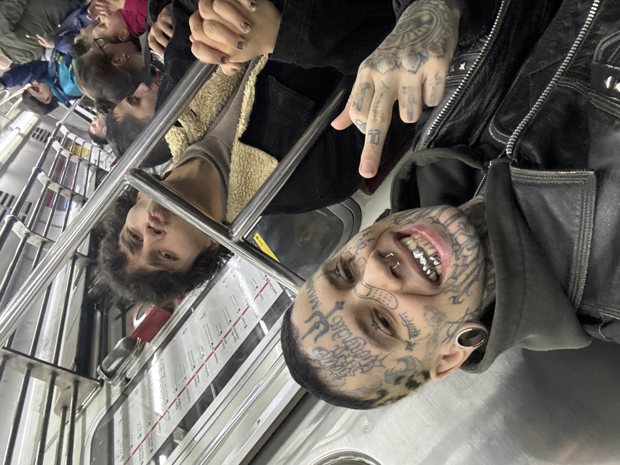 A medium shot of a subway train in Buenos Aires, Argentina, showing a tattooed man with silver teeth grill and large ear gauges. He is wearing a black jacket and gesturing with a tattooed hand. Other passengers are visible in the background, including a person with curly hair reflected in a metal partition with a subway map attached to it. The train car has white walls and metal railings.