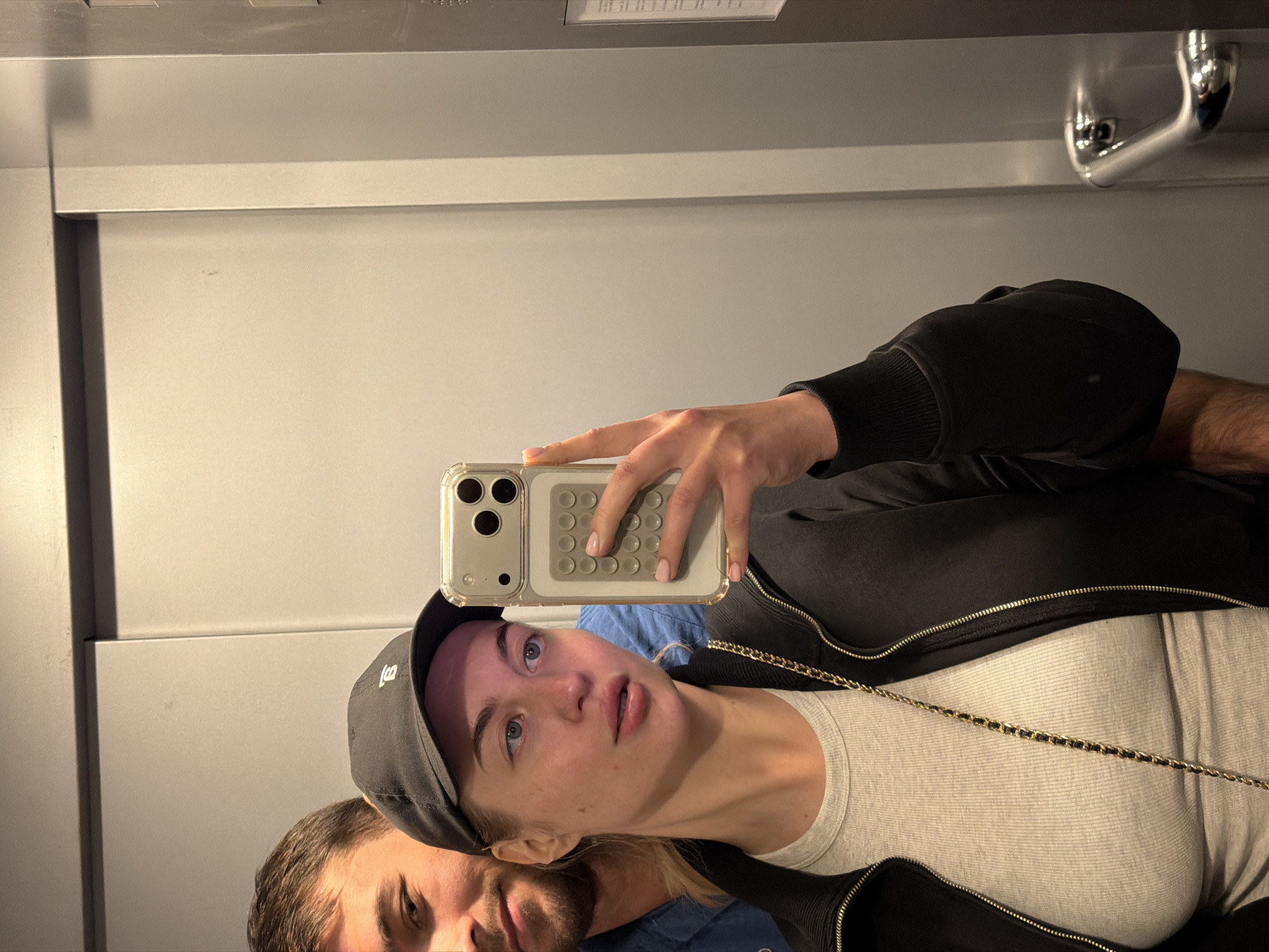This image is a mirror selfie, originally rotated 90 degrees clockwise, showing a woman and a partially visible man indoors. The setting is a minimalist room with light grey walls and reflective surfaces.
The main subject is a woman, depicted from the chest up. She has light-colored skin, light eyes, and light hair that is partially covered by a black baseball cap. She is wearing a black zippered jacket over a light-colored, ribbed top. A thin gold and black chain, possibly from a bag or necklace, is draped across her chest. She holds a smartphone with a prominent triple-lens camera array and a clear case featuring a textured grip on its back, in her left hand, taking the selfie. Her expression is neutral, appearing to look directly into the camera lens. The black baseball cap has a small, unidentifiable white logo on its front.
Behind and slightly below the woman, reflected in the mirror, is a man's face and upper shoulder. He has short brown hair and a beard, and he is wearing a blue shirt. His expression is also neutral.
The background consists of plain light grey walls, with a horizontal structural line. In the upper right corner of the reflected image, a chrome U-shaped pipe fixture, typical of plumbing, is visible. Above the main reflective surface, what appears to be a light fixture or vent is reflected. The lighting is artificial and provides even illumination.
The activity taking place is the woman capturing a mirror selfie with the man present. No specific time of day or weather can be determined from the image. The overall mood is casual. The location context for this image is Sofiivska Borshchahivka, Ukraine. No discernible text is visible beyond the indistinct logo on the cap.