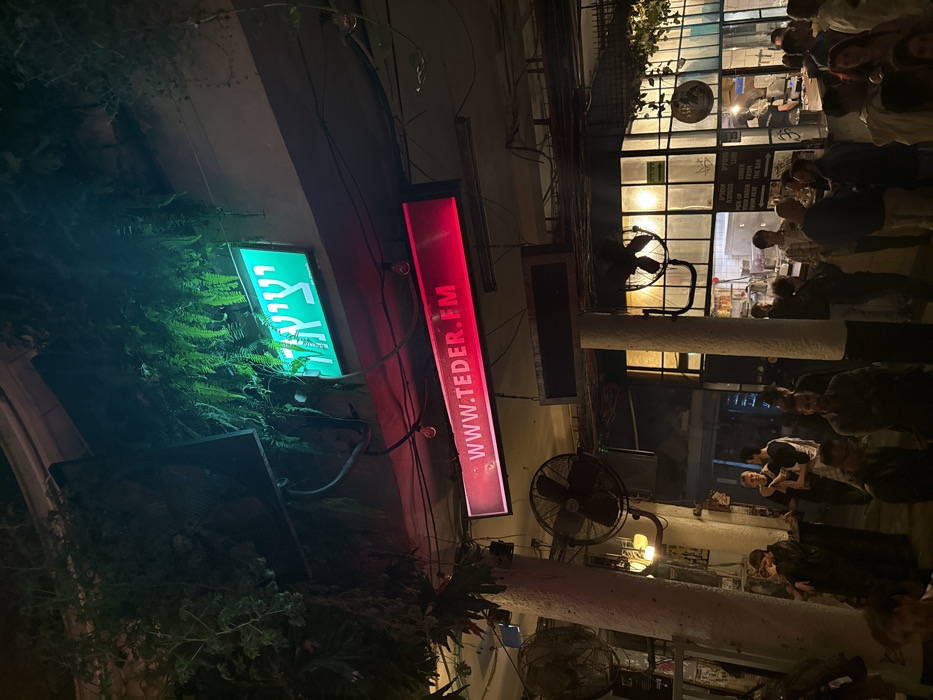 At night, a crowd gathers outside a building with a lit window. A green sign in Hebrew and a red sign reading "www.teder.fm" are attached to the building's exterior, partially obscured by greenery. Several black fans are mounted on the columns supporting the building's structure. The image is captured from a tilted perspective.