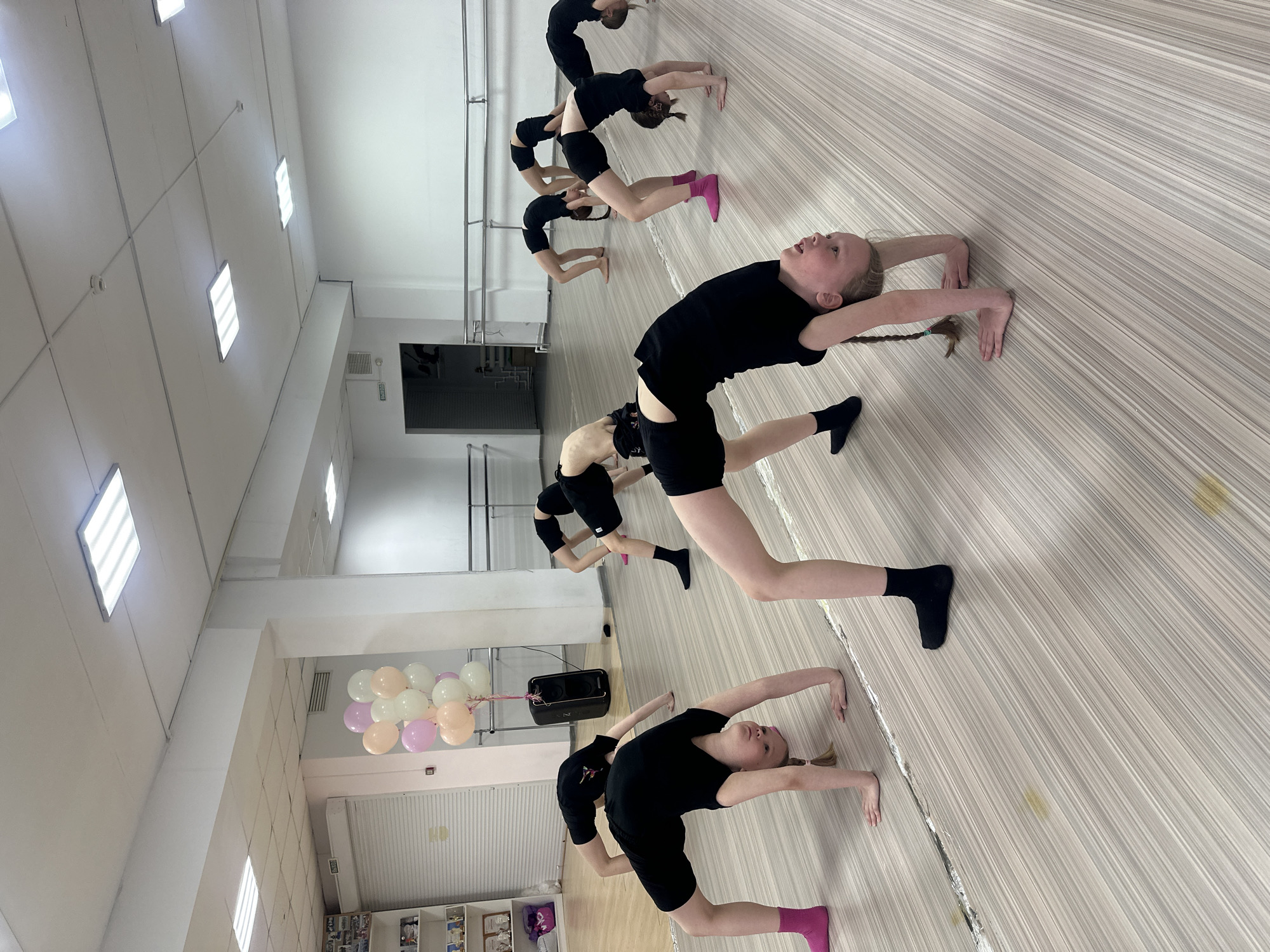 The image shows a group of young girls in a dance studio in what appears to be Votkinsk, Russia, based on the visible architectural features. They are dressed in black dance attire, performing a backbend exercise on a light-colored, wood-patterned floor. There are seven girls visible in various stages of the exercise, each bent backwards with hands and feet on the floor. The studio has white walls, fluorescent lighting, and a mirrored wall with a ballet barre. In the background, there are balloons tied to a speaker and a shelving unit with various items. There is also a closed security gate.