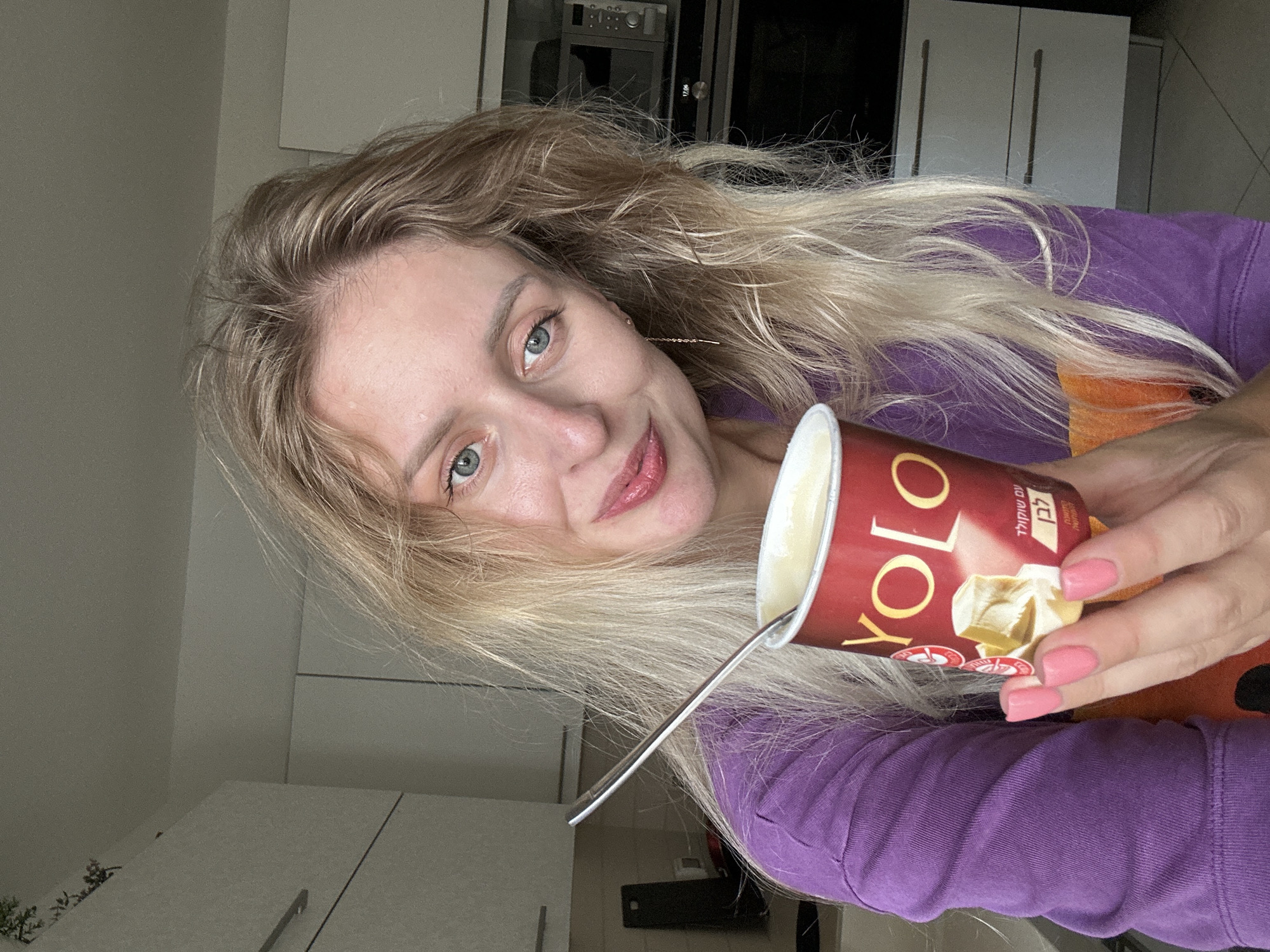 A blonde woman with blue eyes and light skin is holding a red yogurt cup with a spoon in it, in what appears to be a kitchen in Petah Tikva, Israel. She is wearing a purple sweatshirt. The yogurt cup has the brand name "YOLO" and images of white chocolate on it. Behind her, white kitchen cabinets and appliances, including a microwave and oven, are visible. The lighting suggests it is daytime.