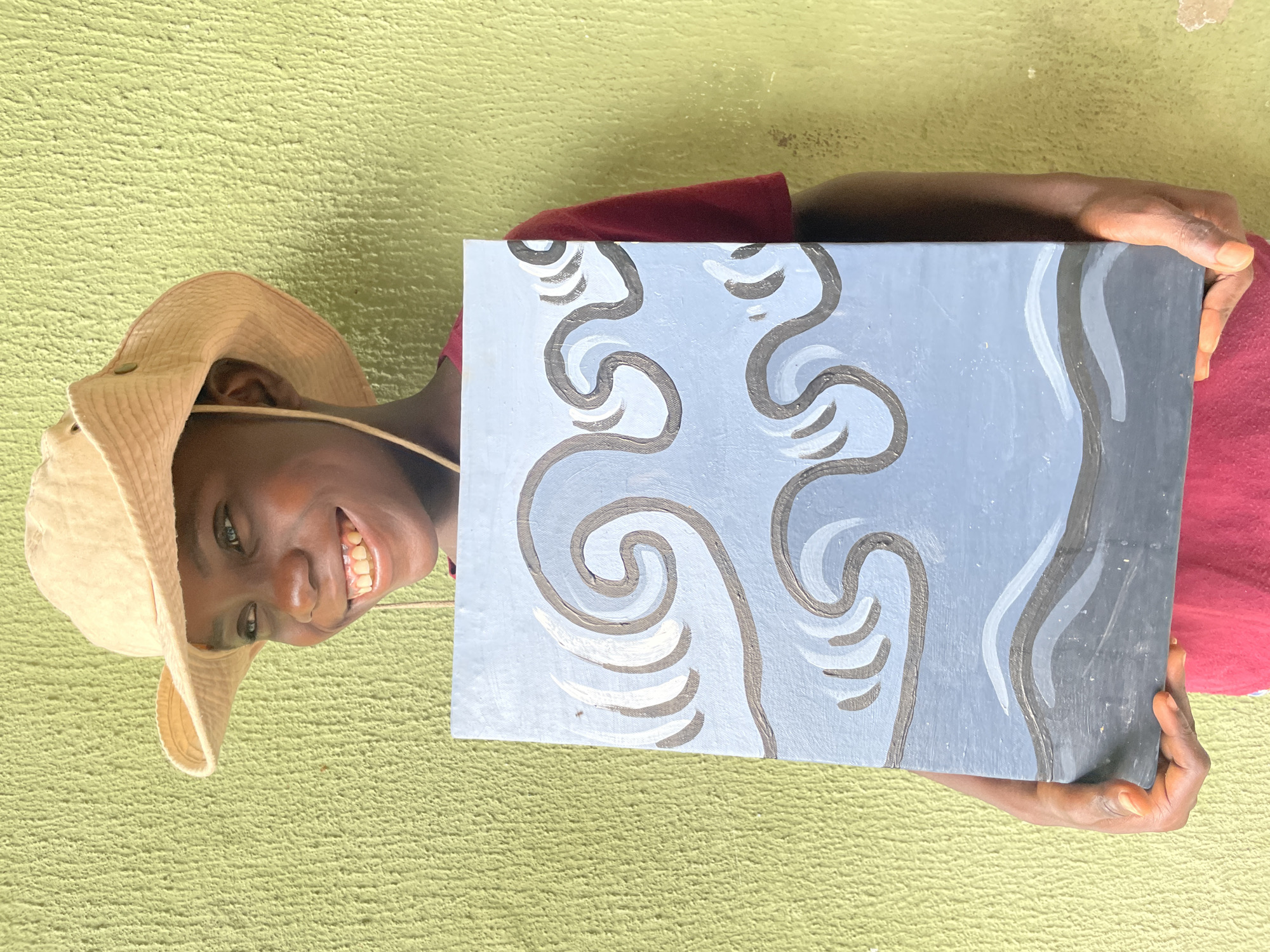 A young Black person, wearing a light-colored bucket hat and a red shirt, smiles broadly while holding up a painting of blue waves against a dark gray foreground. The person's eyes are looking to the left. The background is a textured, light green wall. The image is an outdoor shot, likely in Ado, Nigeria, given the lighting and wall texture, suggestive of a typical residential or public space in the city. The focus is on the person and their artwork, with the environment providing context.