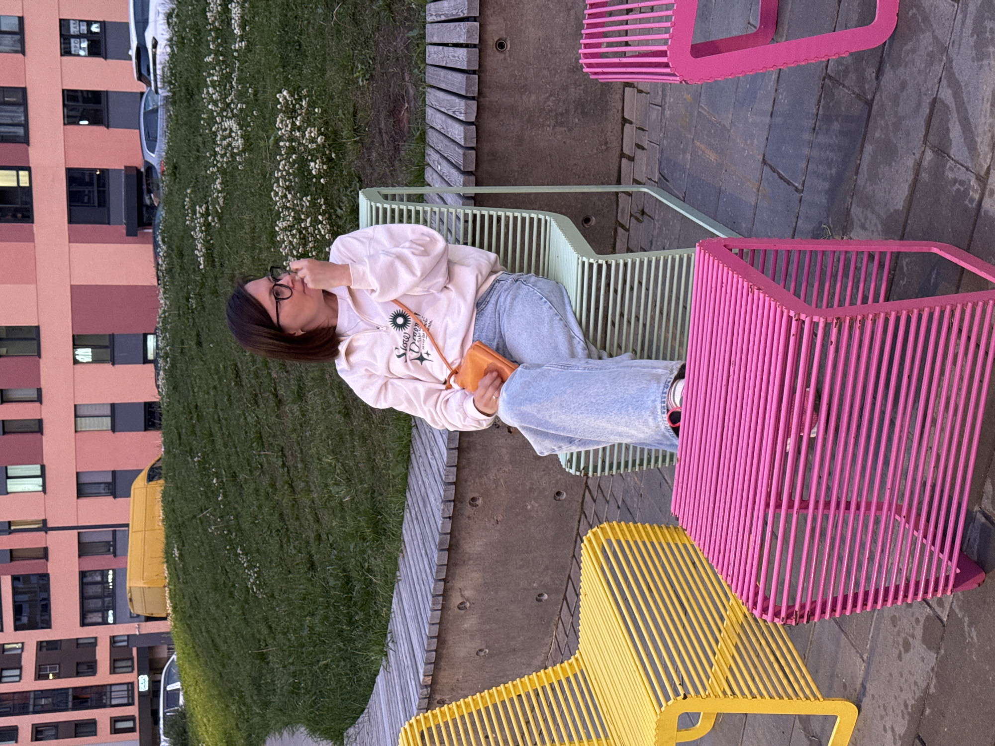 A woman wearing glasses and a light-colored sweatshirt with a graphic print sits on a pale green slatted chair. She has light brown hair and is holding an orange purse on her lap. She wears blue jeans and pink sneakers. In front of her is a bright pink slatted bench. To her left is a bright yellow slatted chair. To her right is another pink slatted bench. The chairs and benches are arranged on a paved area. Behind them, a grassy hill is dotted with small white flowers. In the background, a modern, multi-story building with pink and gray sections is visible. A yellow van and several cars are parked on the street in front of the building. The scene appears to be an outdoor public space in the city of Izhevsk, Russia.