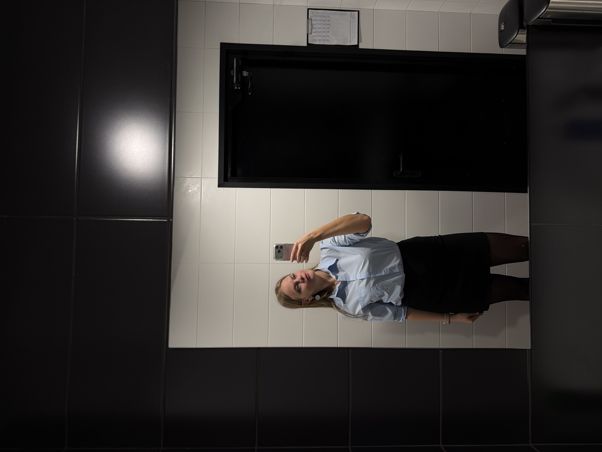 The image shows a woman taking a selfie in a bathroom mirror. The bathroom features white tiled walls with a section of black tiled wall on the left and right sides of the mirror. A black-framed access panel is above the woman, along with a paper sheet attached to the wall. A paper towel dispenser is visible on the right side. The woman is wearing a light blue button-down shirt, a black skirt, and dark tights.
There is no information in the image to indicate the location in Izhevsk, Russia.