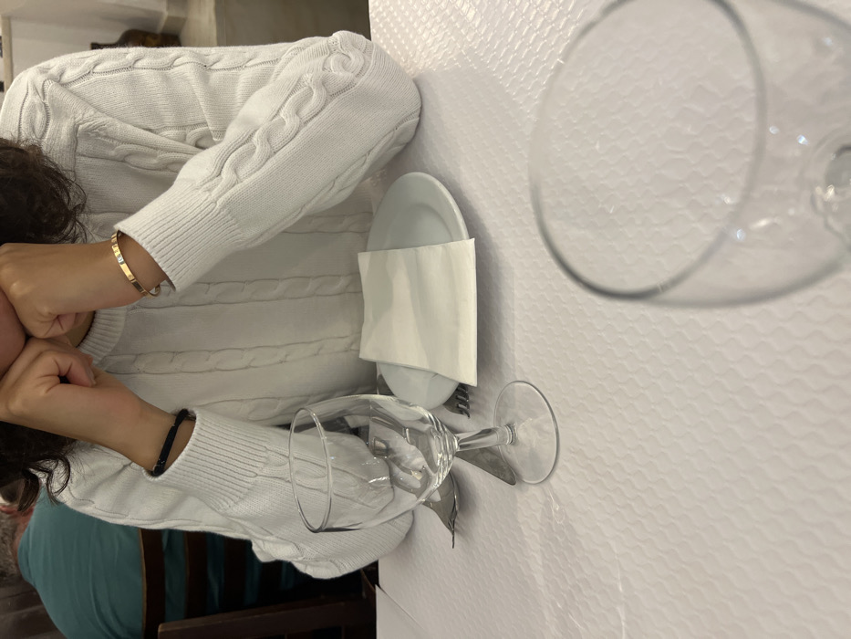 The image depicts an indoor dining scene in Coruche, Portugal. In the foreground, a person wearing a white cable-knit sweater is seated at a table covered by a white textured tablecloth. Their hands are clasped near their lower face. A black cord bracelet is on their left wrist, and a gold-toned bangle with a single square stone is on their right wrist. The table setting includes a white ceramic plate with a folded white paper napkin, a metal fork, and a knife. A clear stemmed wine glass is positioned left of the plate. The rim of a second clear glass is partially visible in the immediate foreground. In the blurred background, a second individual wearing a teal shirt is visible, along with wooden dining chairs and light-colored walls, indicative of a restaurant setting.