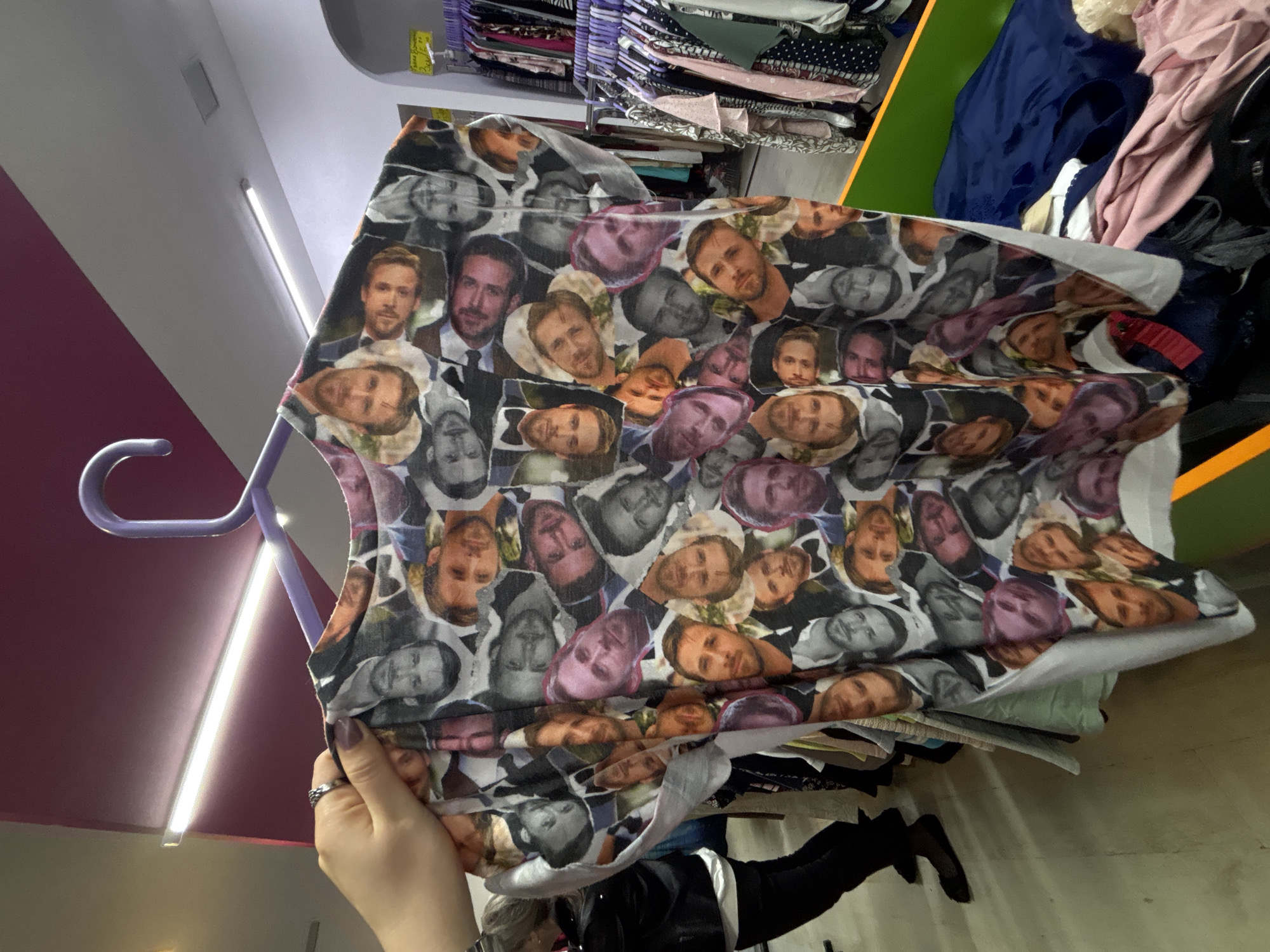 A hand is holding up a t-shirt on a hanger, the t-shirt covered in images of Ryan Gosling. The location appears to be inside a shop with clothing racks and organized shelves. Based on the context of the request including the city of Varna, Bulgaria, this may be a shop located in that area. Details regarding the people include a hand holding the clothing with a silver ring on a finger. The background contains clothing racks, shelves with clothes. The subject of the t-shirt is an illustration of Ryan Gosling's face. The environment is a retail setting.
