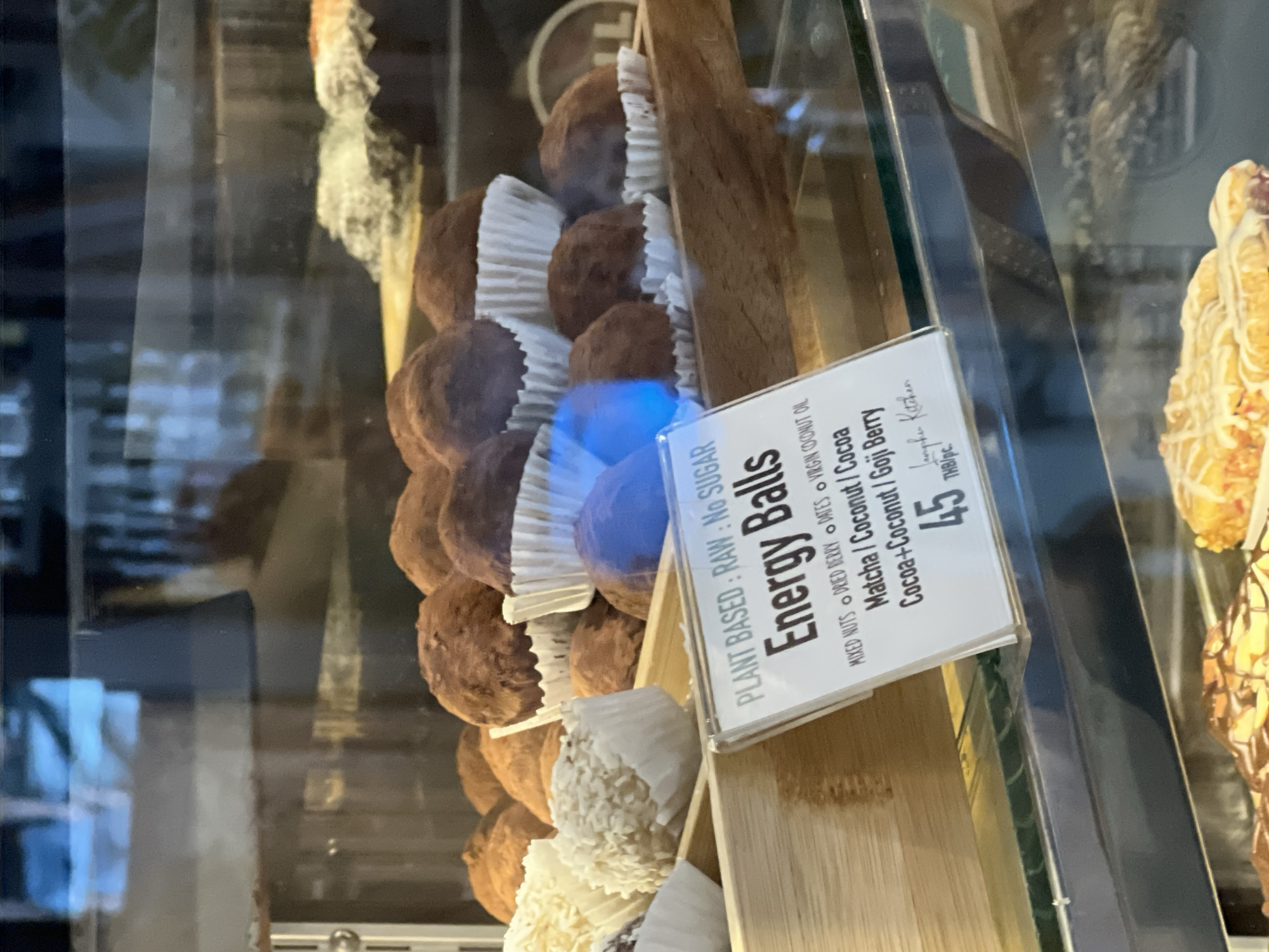 A close-up shot captures an assortment of "Energy Balls" displayed in a glass counter, likely within a cafe or bakery setting in Baan Lamai, Thailand. The main focus is a collection of round, dark brown energy balls, each placed in a white fluted paper liner, arranged on a light-colored wooden tray. One of the balls in the middle of the tray exhibits a prominent blue reflection, possibly from an overhead light or internal display lighting. Towards the lower left, several lighter-colored balls, appearing to be coated in coconut, are also visible in similar paper liners.
A white informational sign is prominently displayed in front of the energy balls. The text on the sign details the product: "PLANT BASED : RAW : No SUGAR", "Energy Balls". It also lists ingredients and flavor variations such as "MIXED NUTS • DRIED BERRY • DATES • VIRGIN COCONUT OIL", "Matcha / Coconut / Cocoa", and "Cocoa + Coconut / Goji Berry". The price for one piece is clearly stated as "45 THB/pc." (Thai Baht per piece), and the name "Langhe Kitchen" is partially visible at the bottom of the sign. To the right, part of another textured, golden-brown dessert item is visible within the display. The reflections on the glass casing indicate an indoor environment with ambient lighting.