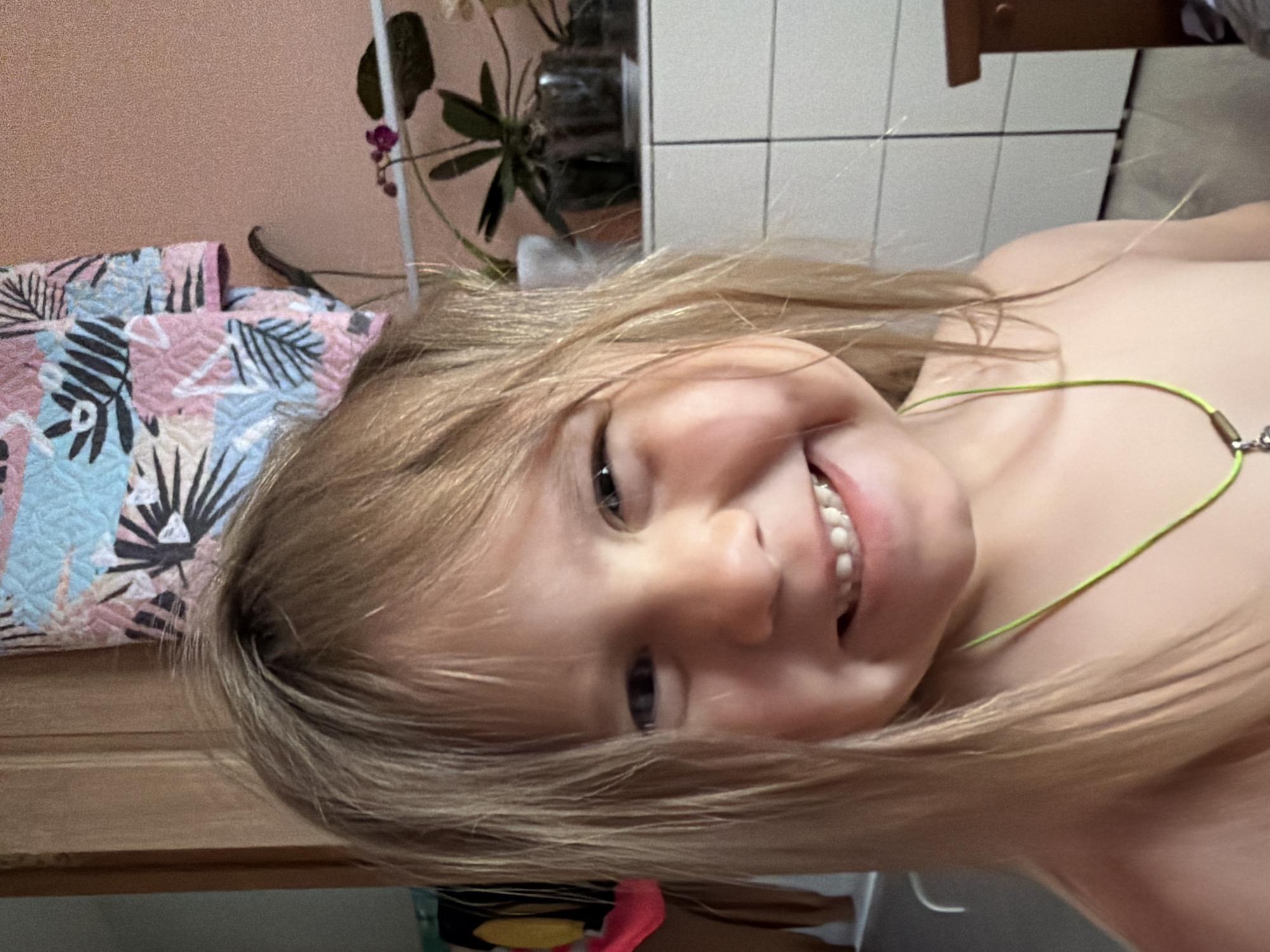 A young child with blonde hair is smiling at the camera. The child is wearing a thin, lime green necklace. Behind the child is a plant with green leaves and purple flowers, a white tiled wall, a wooden cabinet, and a bed with a patterned quilt. It is impossible to confirm the location is Chaikovsky, Russia, from the provided image.