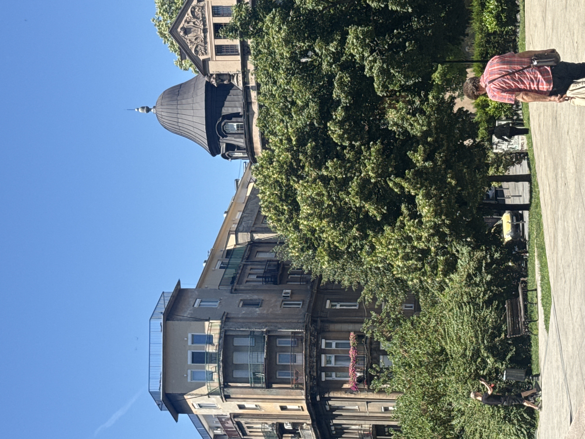 In Belgrade, Serbia, a clear blue sky is visible over a city scene featuring architecture and greenery. A multi-story building with a weathered facade and balconies dominates the left side of the frame, its upper floors featuring windows and railings. To the right, a dark domed structure is partially obscured by lush green trees, with a portion of another building featuring decorative relief visible behind it. In the foreground, a paved walkway runs alongside a grassy area with a park bench and dense foliage. A person in a black t-shirt and shorts walks to the left, seemingly interacting with a trash receptacle, while another person in a red and white plaid shirt walks away from the camera, carrying a bag over their shoulder. The scene captures a sunny day in an urban environment, with a mix of historical buildings and natural elements.