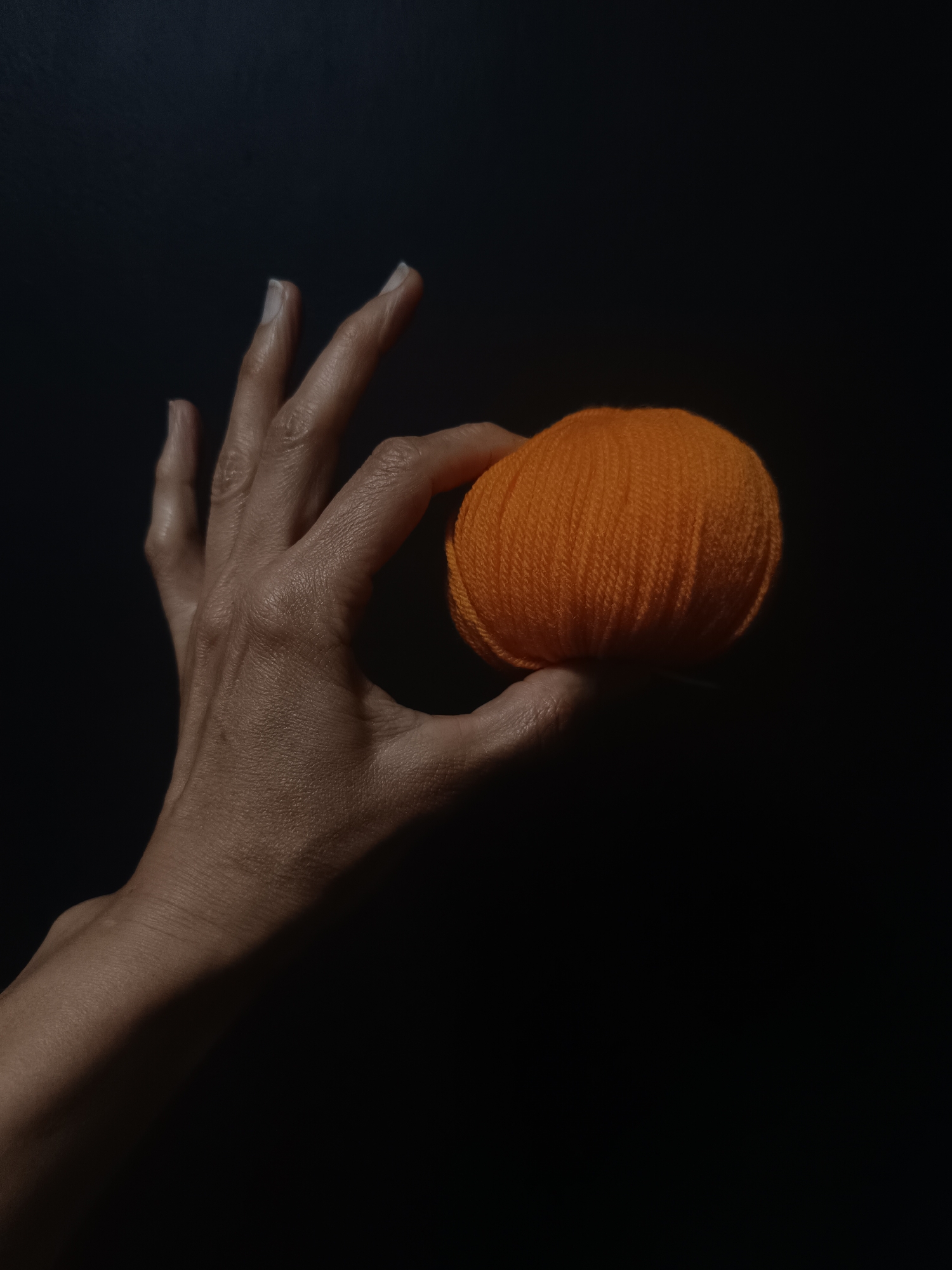 A hand holds a small orange ball of yarn against a dark background. The skin on the hand appears textured, and the nails are long. No information suggests the scene is in Lermontov, Russia.