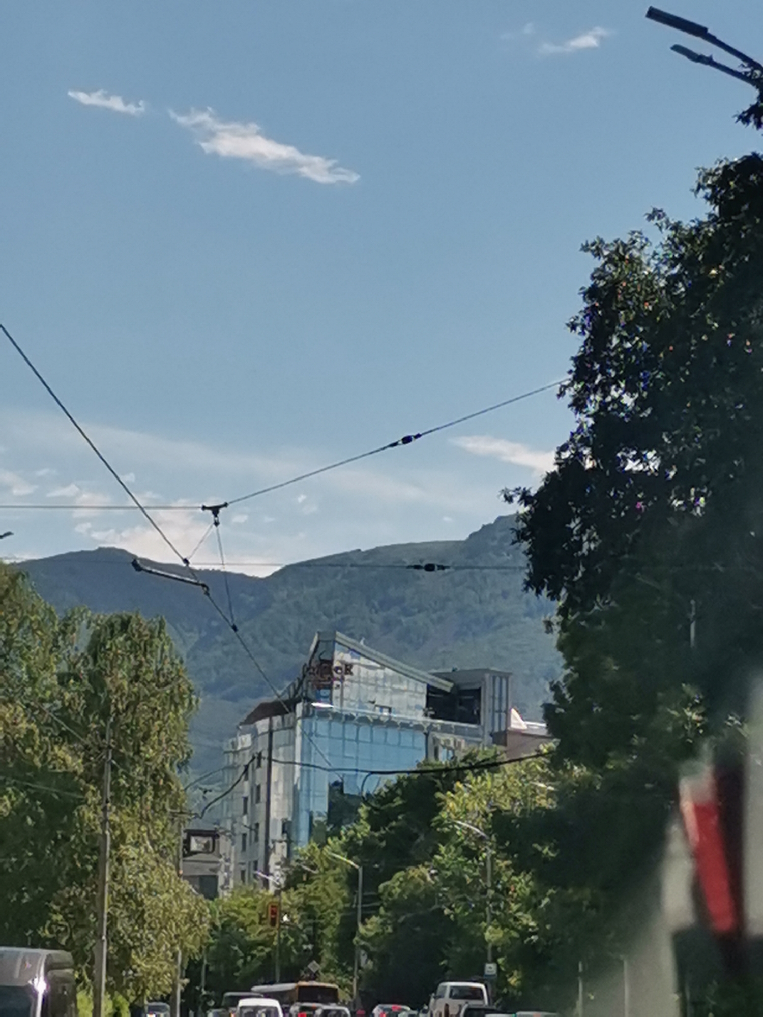 The image shows a view of a city street in Sofia, Bulgaria, with a modern building in the background. The building has a glass facade and appears to be an office building or hotel. The street is lined with trees, and there are several cars and a bus on the road, indicating moderate traffic. Overhead wires, likely for public transport, crisscross the sky. In the distance, a mountain range is visible, its slopes covered in green vegetation. The sky is blue with a few wispy clouds. The overall impression is that of an urban environment on a sunny day.
