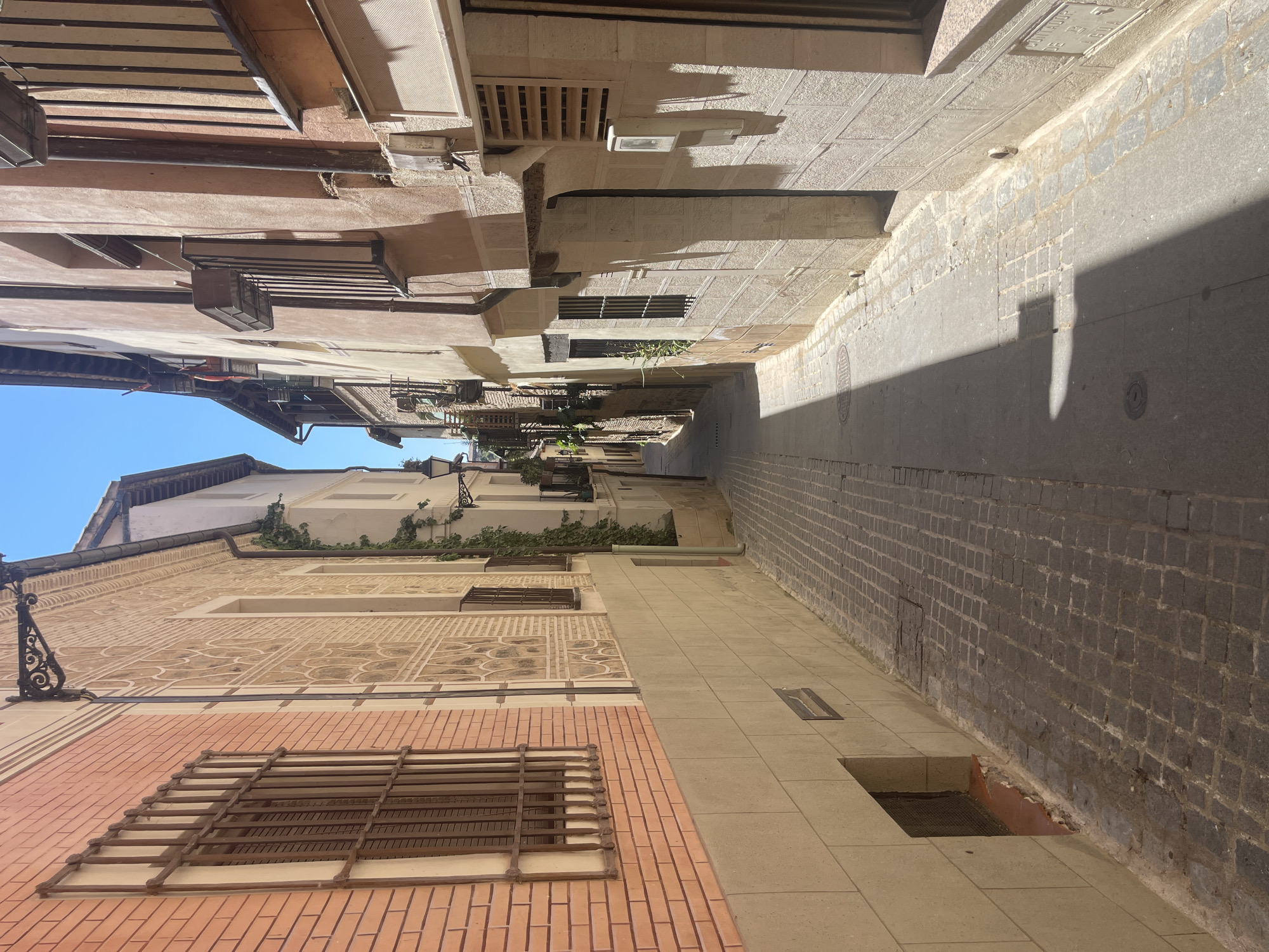 This is a vertical, eye-level, outdoor photograph taken on a sunny day in Toledo, Spain. The image depicts a narrow cobblestone street lined with old buildings. The street curves slightly to the right and recedes into the distance, where it becomes obscured by shadows.
On the left side of the street, there is a building with a salmon-pink brick facade on the lower section and a cream-colored upper section. The upper section has a decorative patterned frieze. A window with a brown metal grate is visible on the brick section. There are small, dark vents or openings near the base of this building.
The building on the right is constructed with rough, beige stone blocks on the lower section and a lighter, stucco-like material on the upper section. This building features several windows, some with dark metal grates and small balconies with black iron railings. There are various architectural details, including drainpipes, a protruding ledge, and electrical boxes mounted on the facade. One of the upper windows has a slatted metal grate.
The cobblestone street is well-worn and shows some signs of age. The sunlight casts a strong, diagonal shadow across the right side of the street, obscuring portions of the buildings and the pavement. The sky above is a clear, bright blue. The scene appears to be an older part of the city, characterized by its narrow streets and historic architecture. There are no people or animals visible in the image.