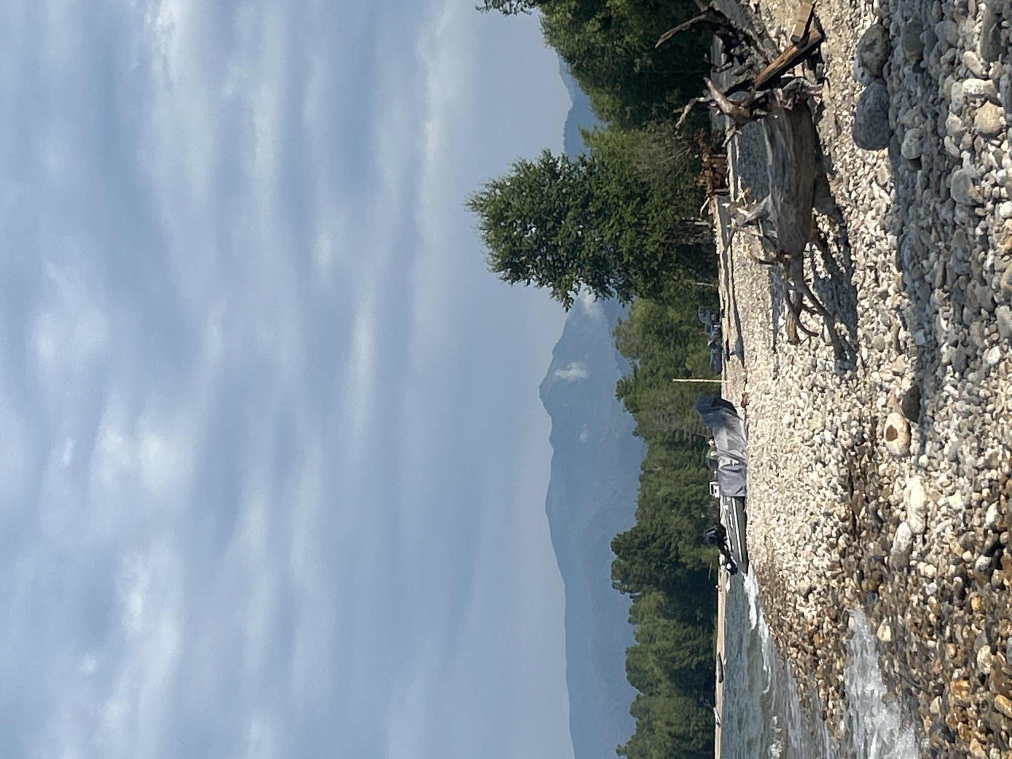 A rocky shore of a body of water is depicted with the water lapping at the stones. In the foreground, a large piece of driftwood rests on the shore. Further back, several inflatable boats with engines are visible, some covered with tarps. A dense forest line runs along the horizon, with mountains visible beyond the trees. The sky is overcast with clouds. This scene is located in the city of Tolbazikha, Russia.