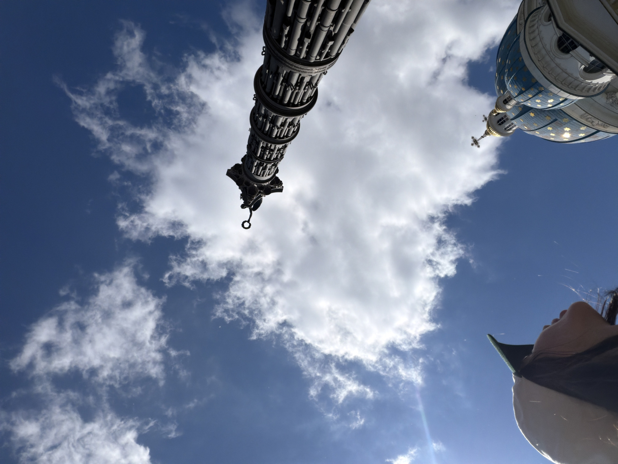The image, captured from a low angle looking upwards, showcases a dramatic sky filled with white, puffy clouds against a vibrant blue backdrop. In the foreground, a young woman wearing a light-colored baseball cap with a green brim is visible, her head tilted back as she gazes upwards. To the right, the ornate blue domes of a Russian Orthodox church, likely the Kazan Cathedral in Saint Petersburg, Russia, are prominently featured, adorned with golden stars and topped with crosses. Further up and to the left, a tall, dark, cylindrical monument or column stands against the sky, appearing to be constructed from multiple stacked sections. The overall impression is one of awe and architectural grandeur set against a bright, cloud-filled sky.